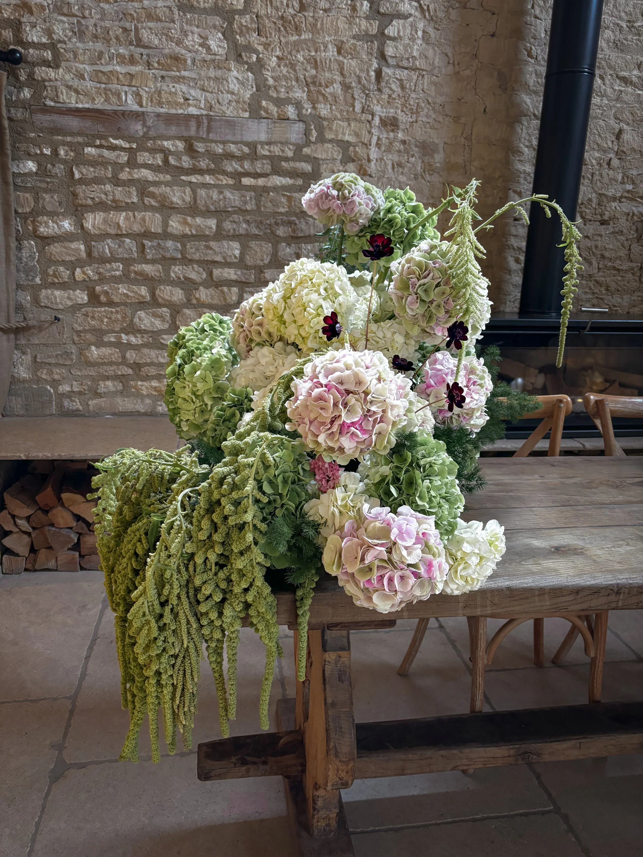 Modern ceremony floral installation at Old Gore Barn in the Cotswolds, Gloucestershire, using hydrangea, draping green amaranthus, soft pink flowers and chocolate cosmos.