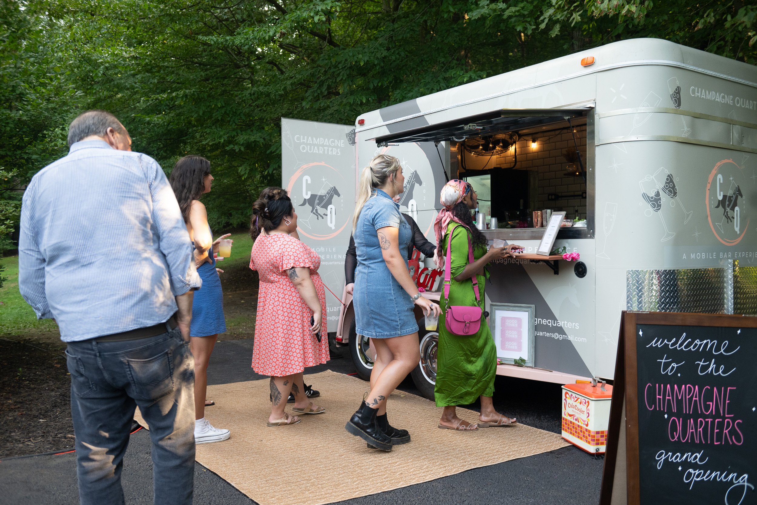 Group of people standing in line at a mobile bar trailer labeled Champagne Quarters in outdoor park area, with trees in the background and a welcome sign on a chalkboard outside the trailer.