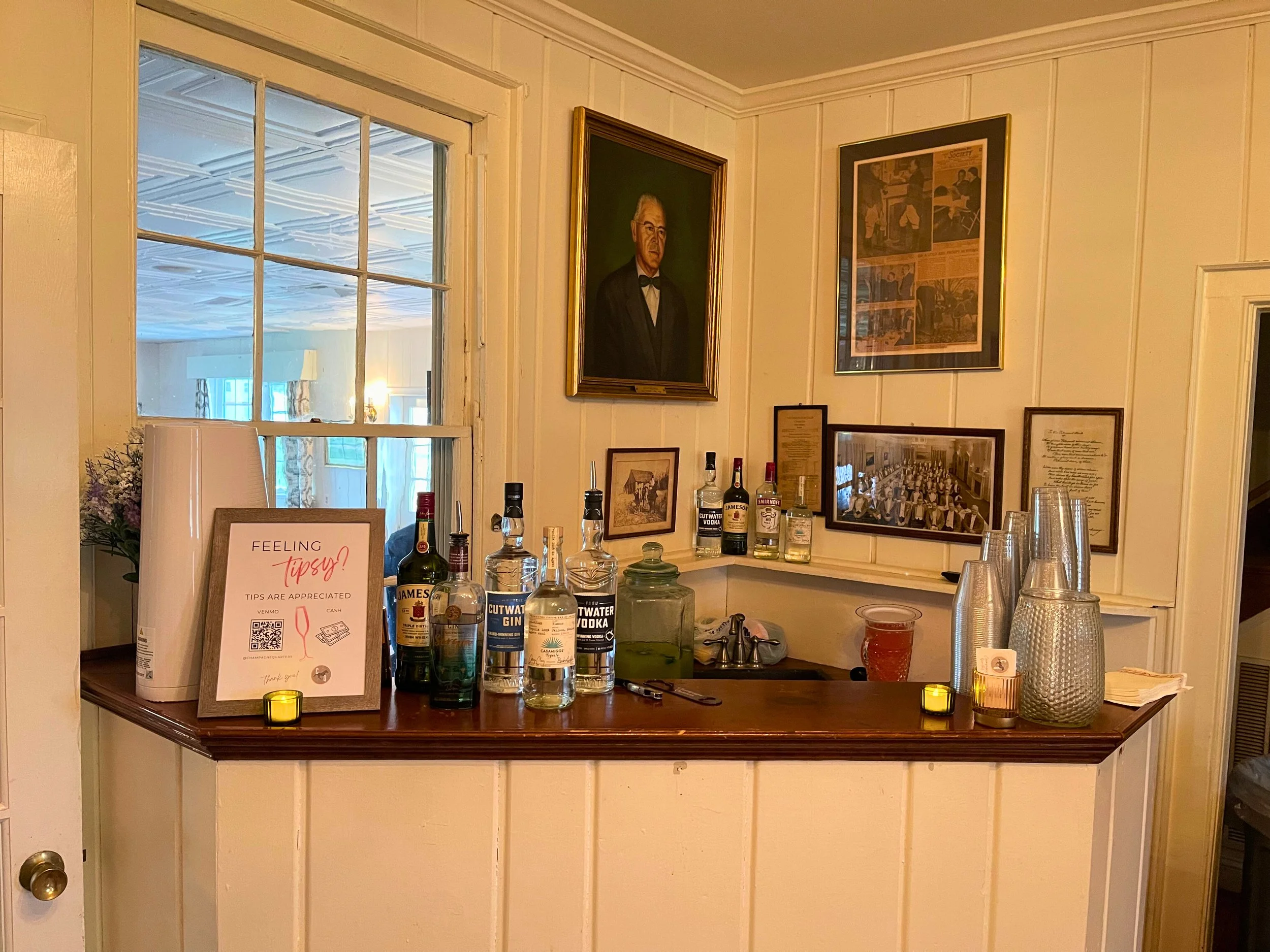 A small home bar area with a variety of liquor bottles, plastic cups, and a sign about tips. The wall behind the bar has framed photographs and a portrait of an older man in a tuxedo. There is a window to the left showing a room with lights and furni