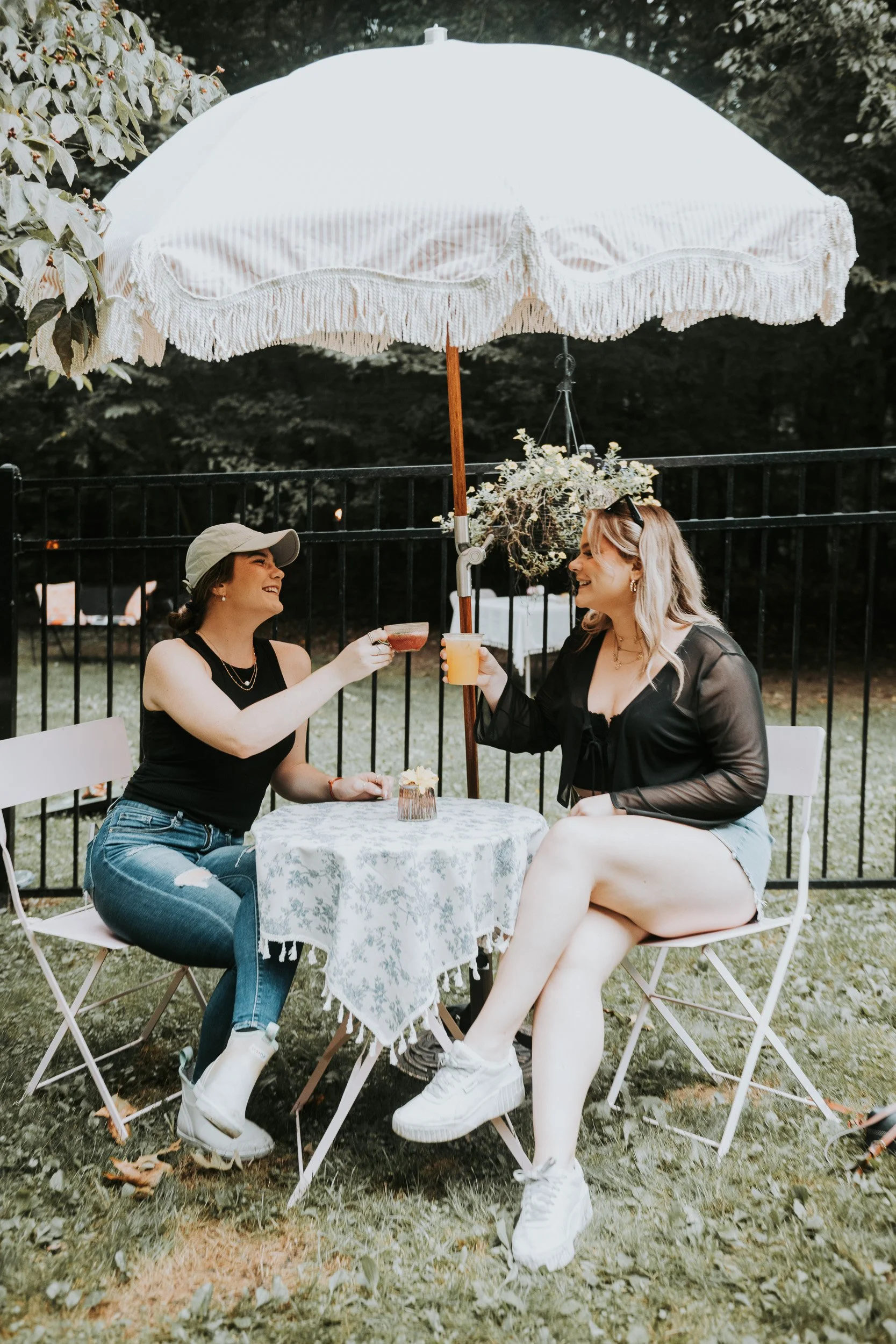 Two women sitting at a small round table under a white patio umbrella, raising drinks and smiling at each other outdoors.