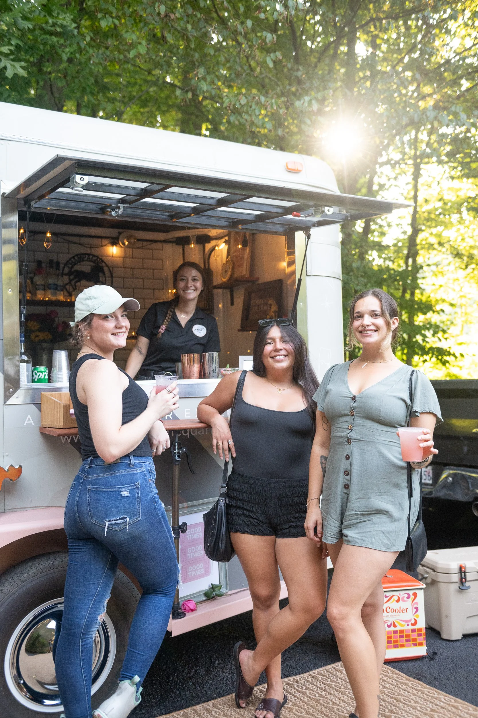 Four women standing outside a food truck, smiling and holding drinks, with a woman inside the truck serving customers. Sunlight filters through trees in the background.