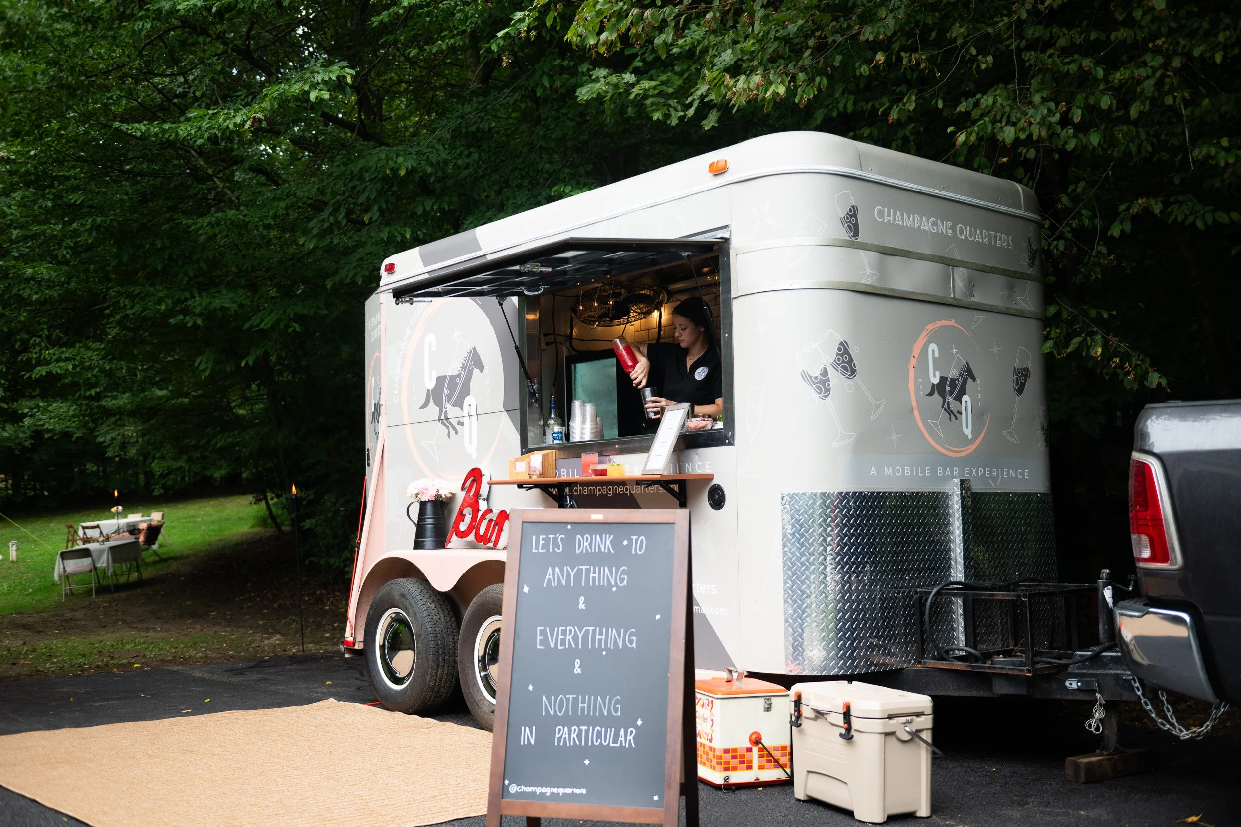 A mobile bar trailer with a bartender pouring a red drink through a shaker, outdoor setting with green trees, a chalkboard sign with the message 'Let's drink to anything & everything & nothing in particular', and a small table with chairs in the back
