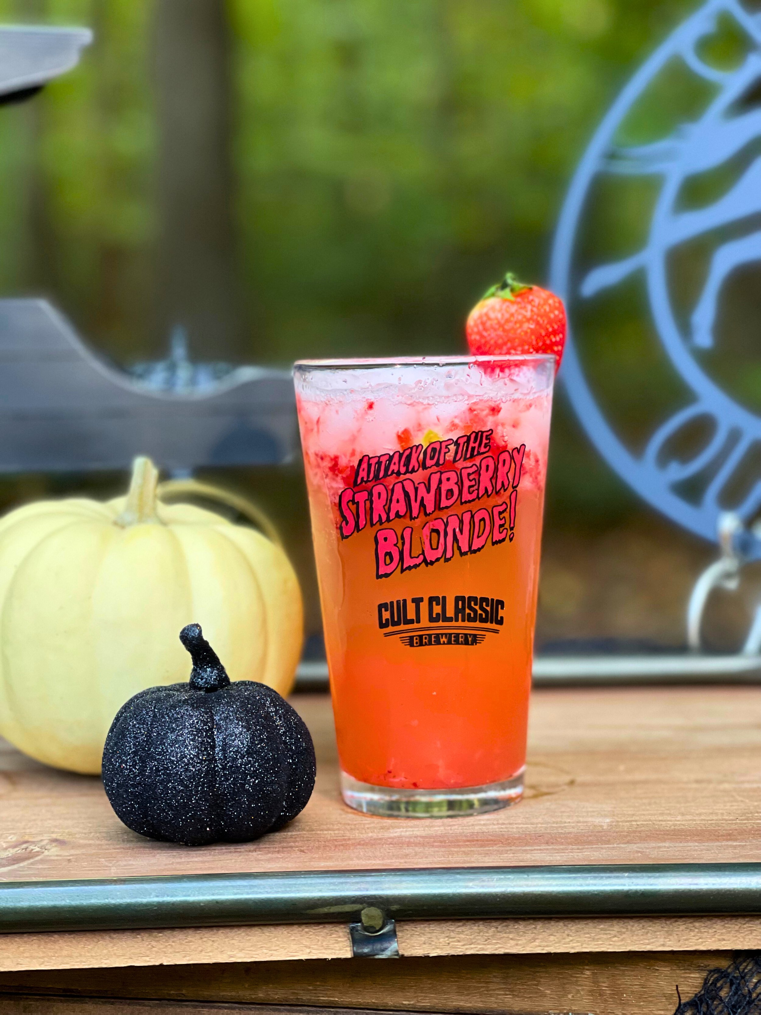 A tall glass containing a pinkish strawberry drink with a strawberry on top. The glass has a colorful label that reads 'Attack of the Strawberry Blonde! Cult Classic Brewery.' On a wooden surface, next to the glass, there is a white pumpkin and a bla