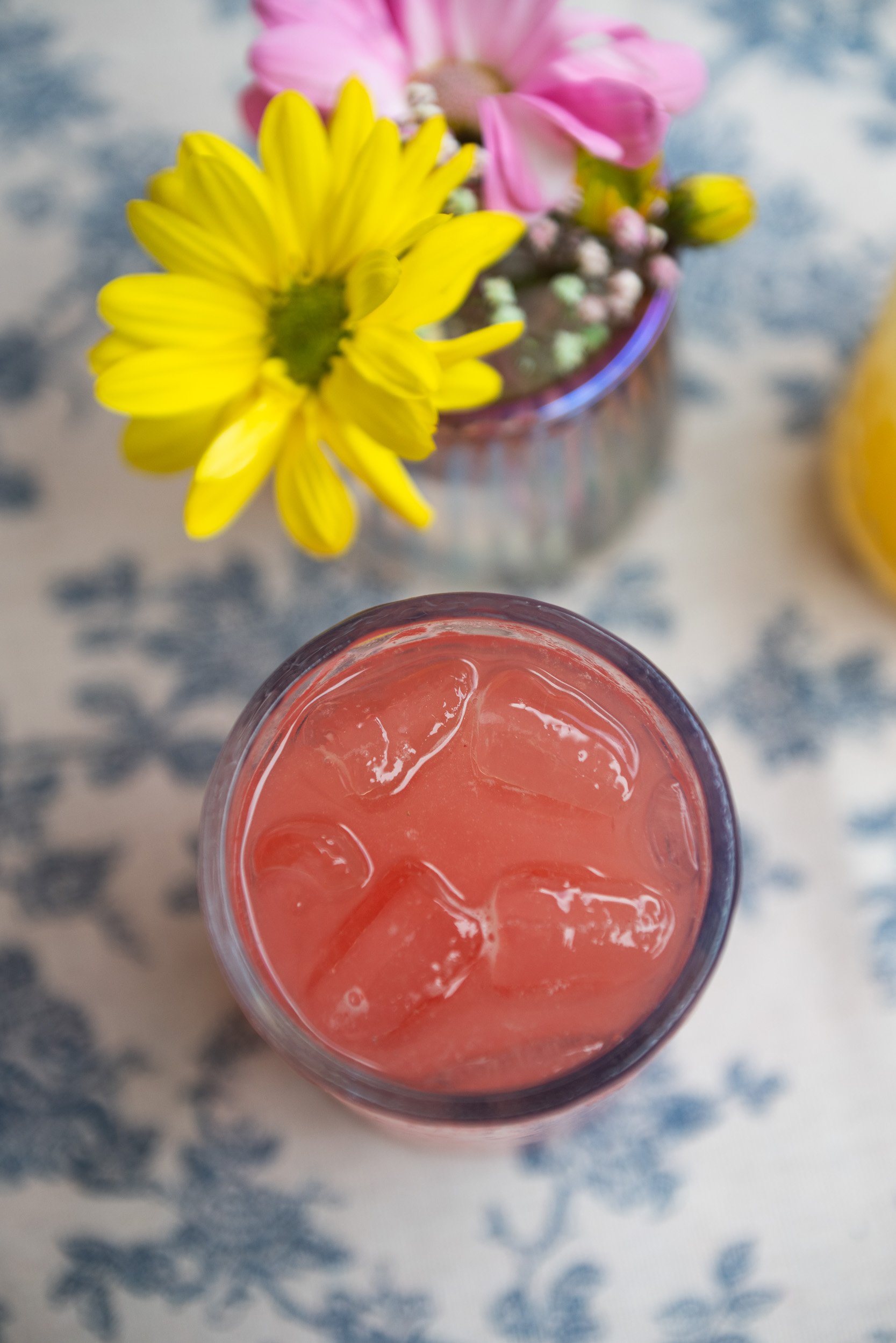 A top-down view of a pink beverage with ice cubes in a glass, placed on a blue and white patterned surface, with a blurred flower arrangement in a glass jar in the background.
