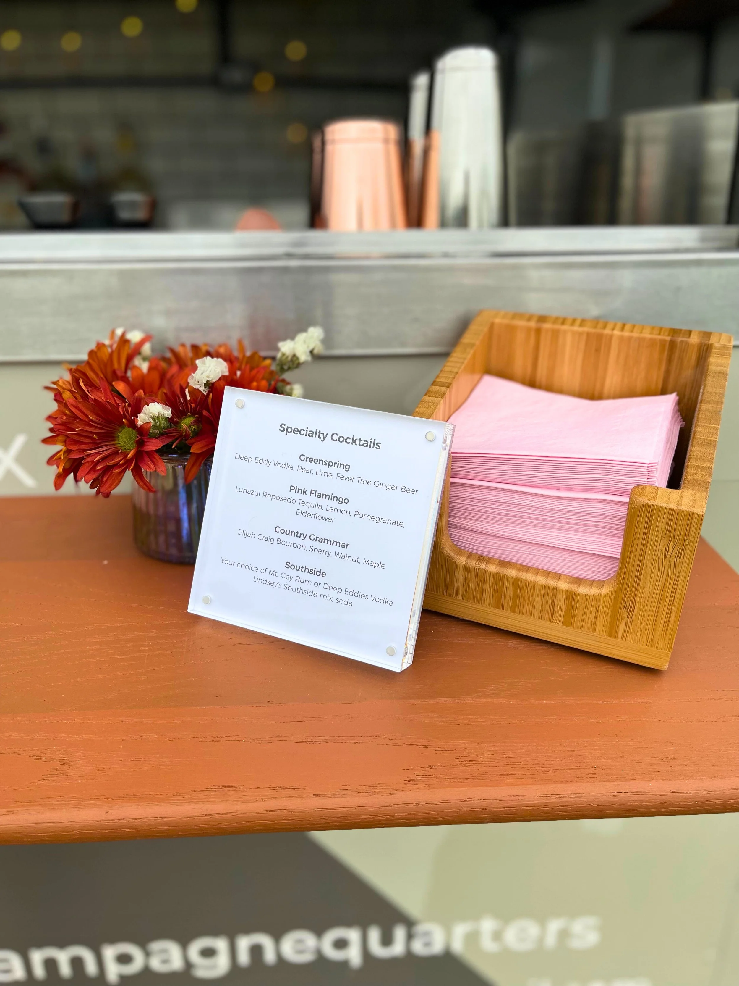 A table displaying a bouquet of red and orange flowers, a sign listing specialty cocktails, a wooden napkin holder with pink napkins, and a blurred background of a bar or kitchen area.