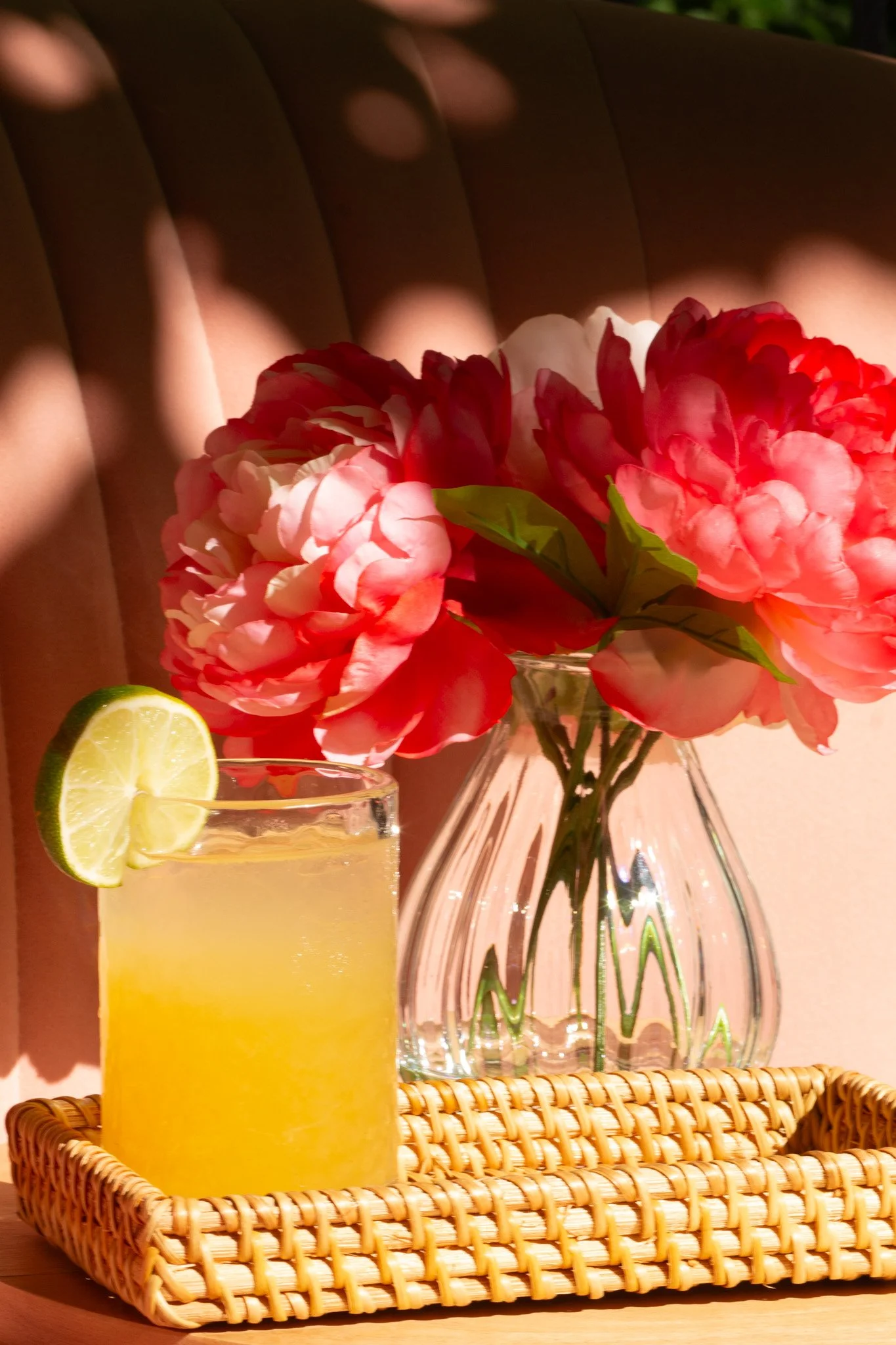 A glass of yellow lemonade with a lime wedge on the rim, placed on a wicker tray, and a vase with pink peony flowers in the background, set against a warmly lit wall with soft shadows.