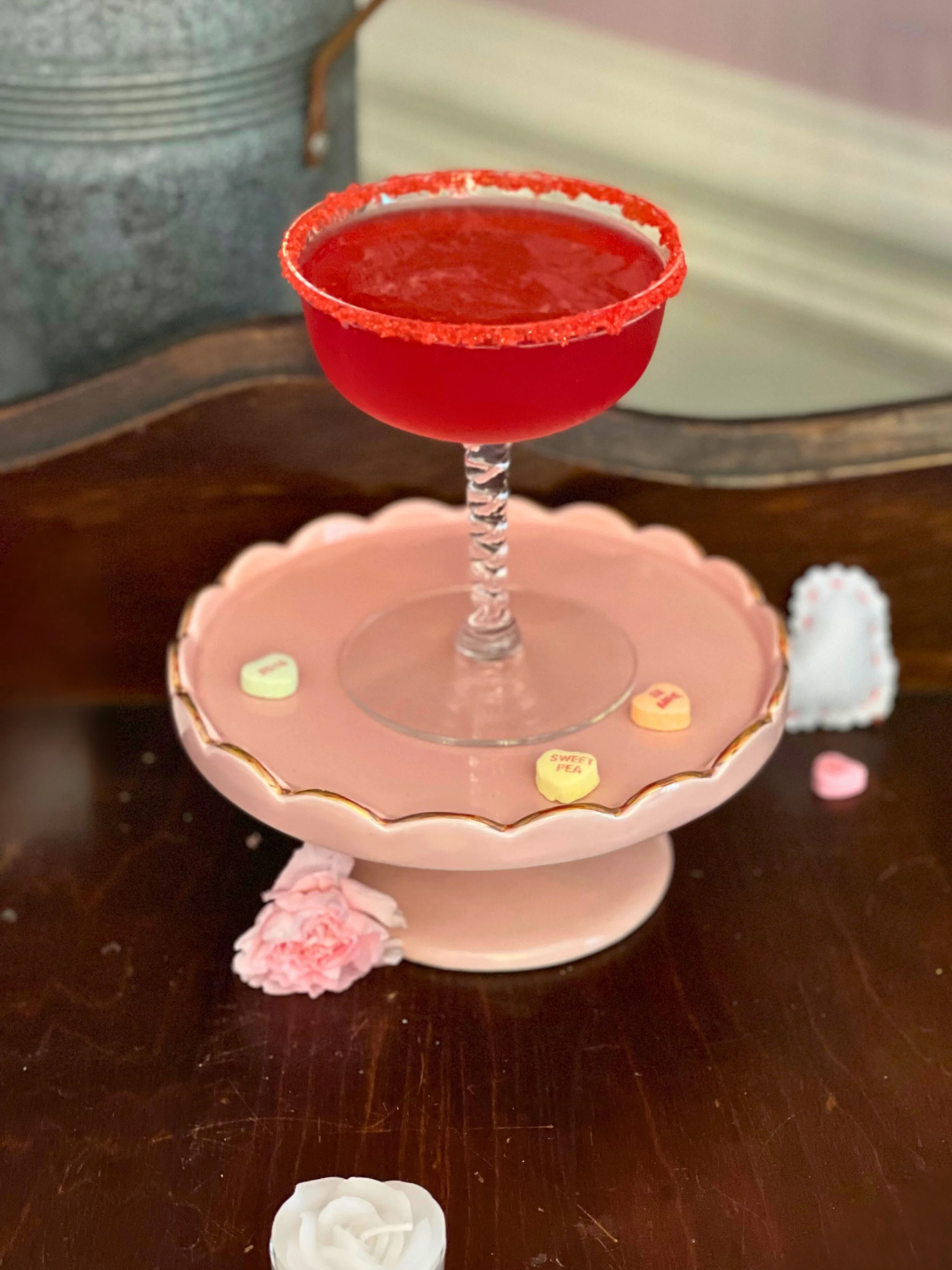 Red cocktail with a salted rim in a coupe glass, placed on a pink decorative cake stand surrounded by small pastel candy hearts and pink flowers.