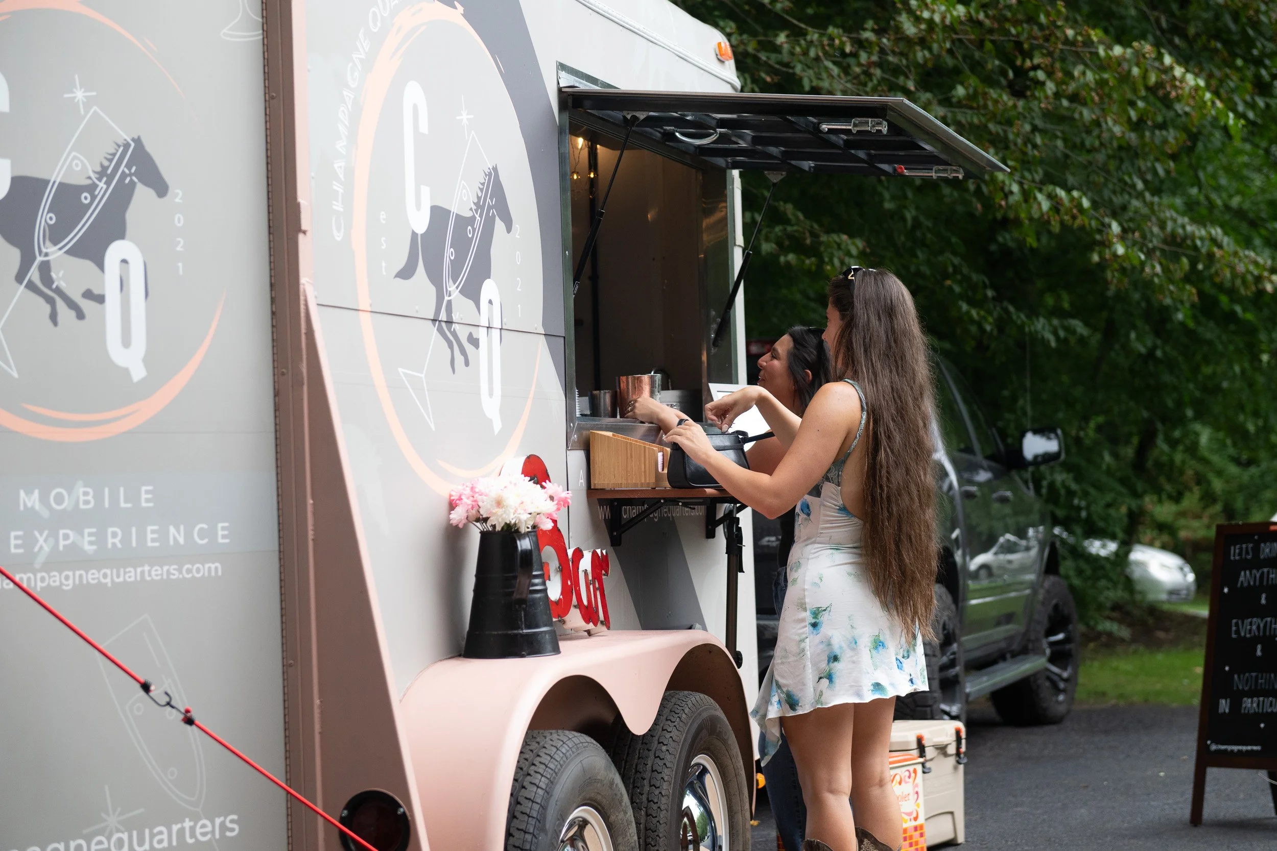 Two women order cocktails from a food truck with space-themed logo, outside on a green, wooded area.