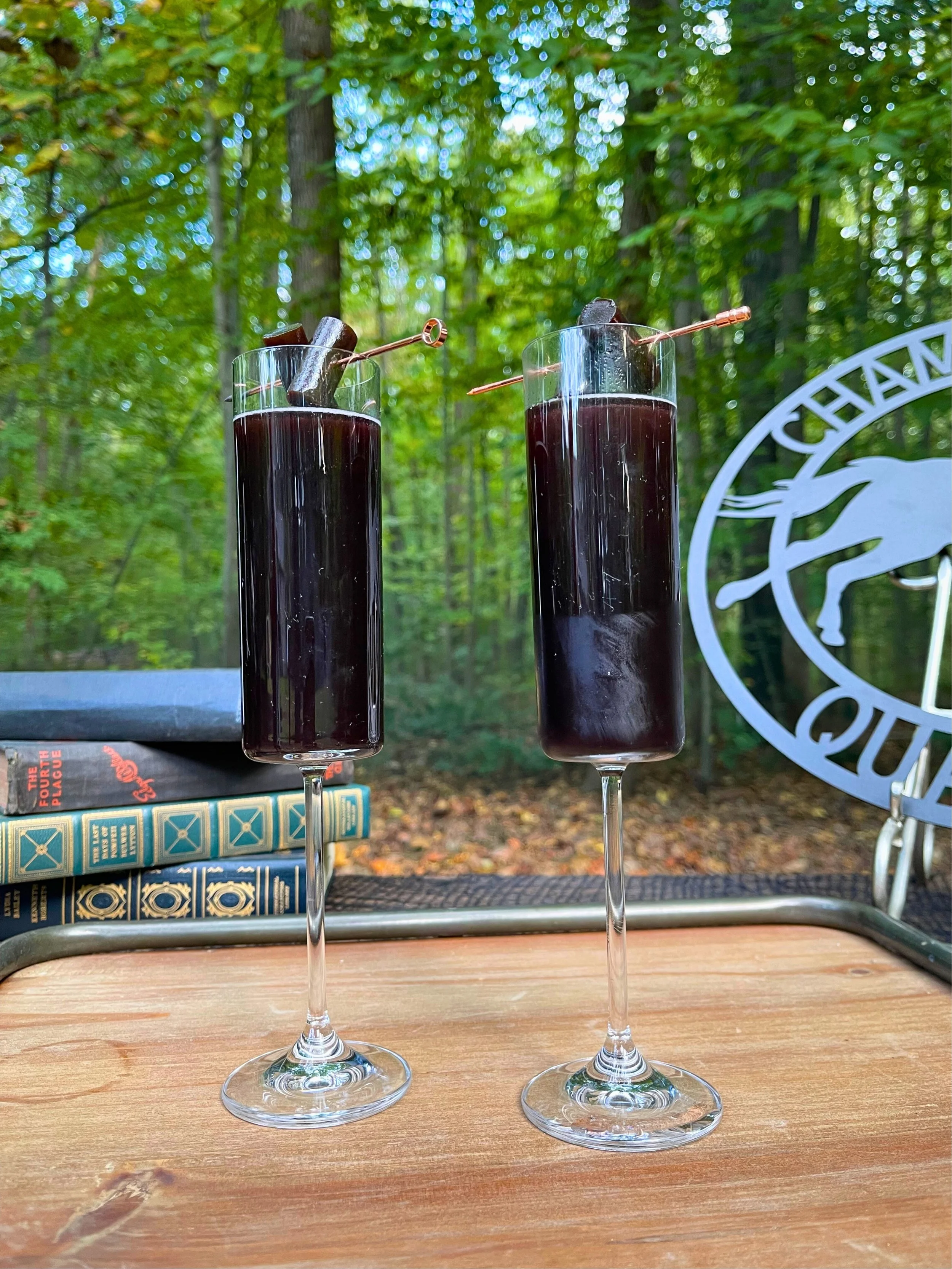 Two tall glasses filled with a dark red or purple beverage, garnished with charcoal and small metal skewers, set on a wooden table outdoors with a forest background.