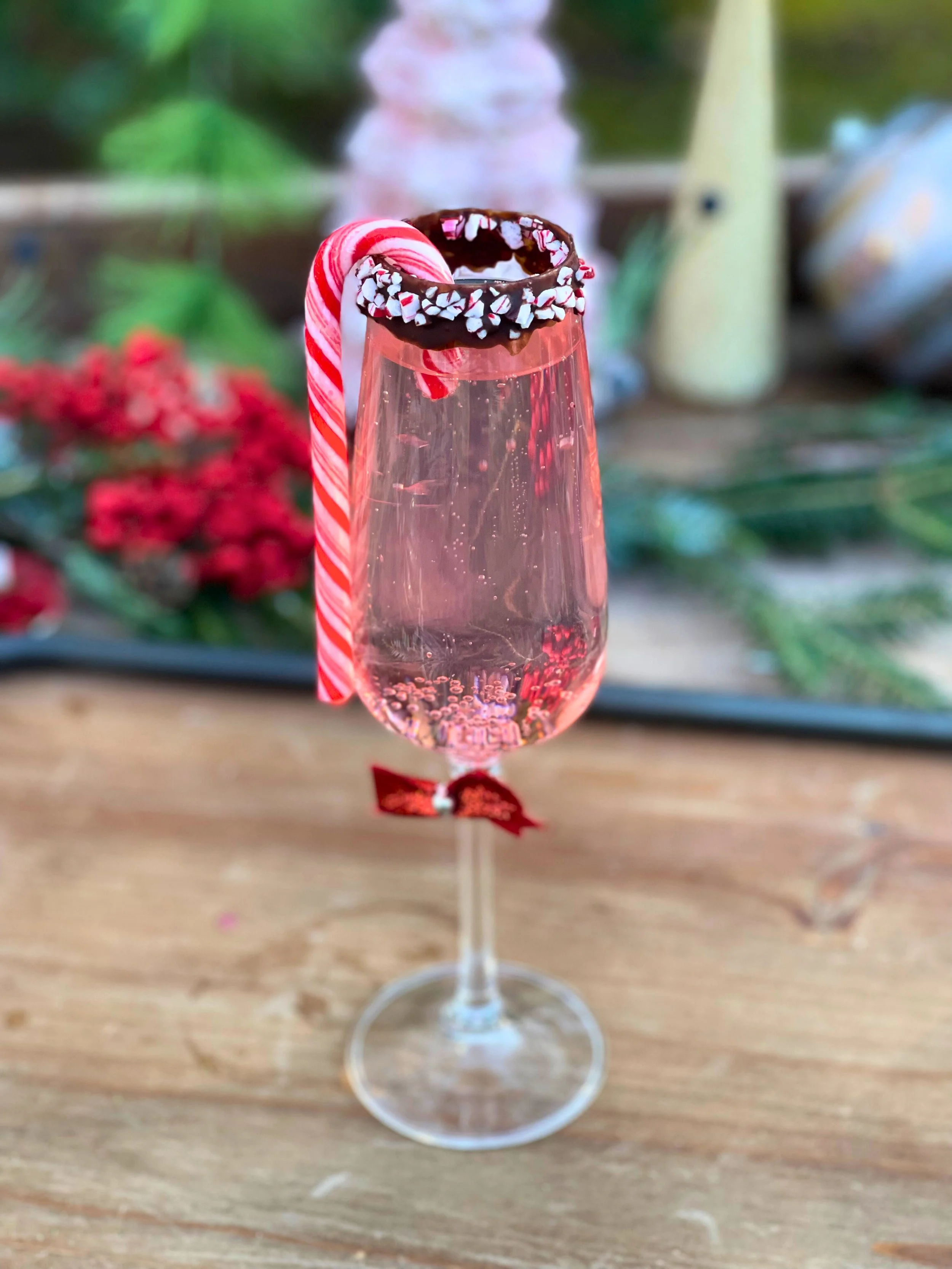 A tall glass filled with pink sparkling drink, topped with a chocolate-dipped candy cane with red and white stripes, a chocolate-covered rim with white sprinkles, and a small red bow tied around the stem.