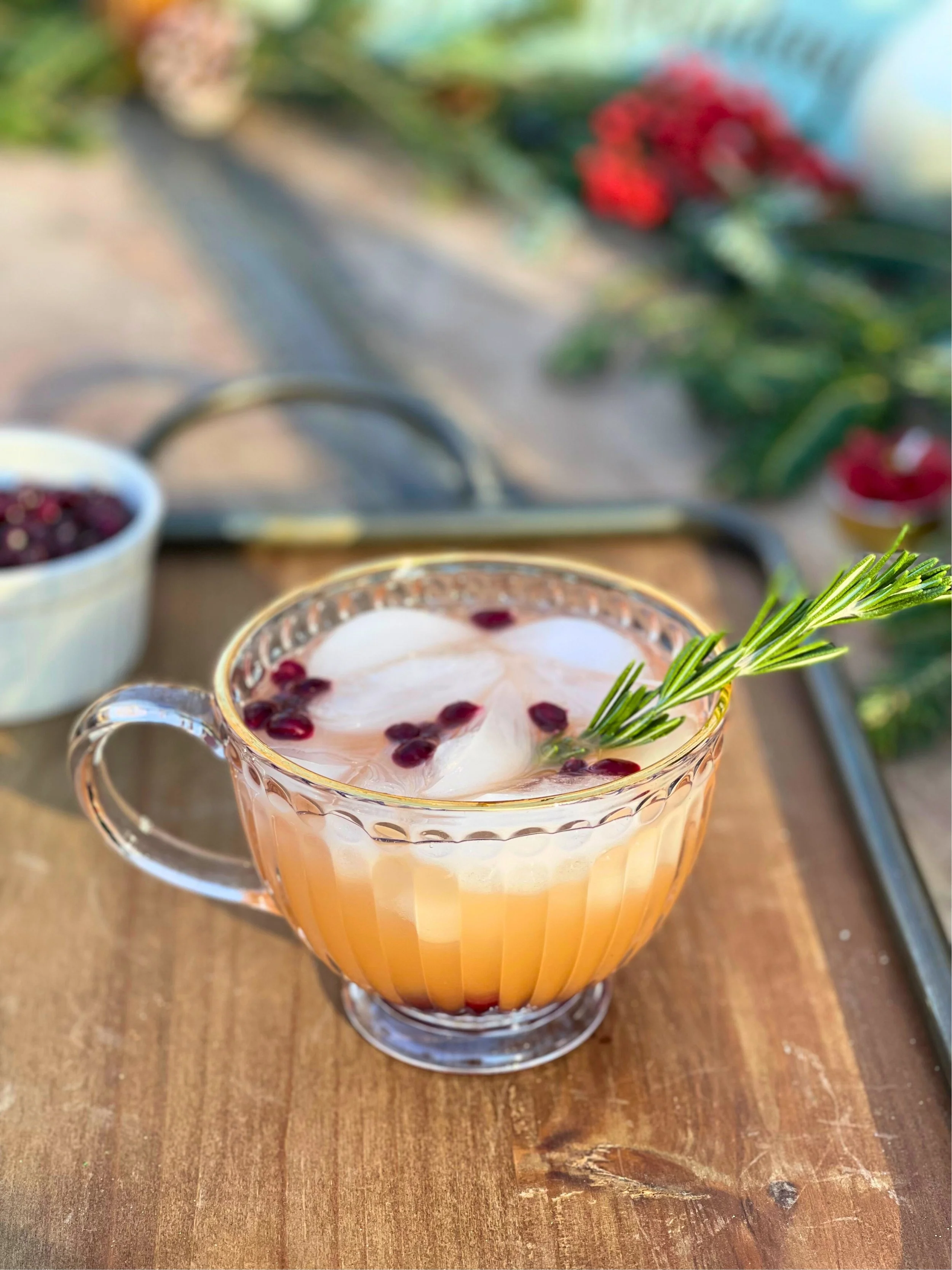 A glass cup of pink lemonade with ice, pomegranate seeds, and a sprig of rosemary on top, placed on a wooden surface.