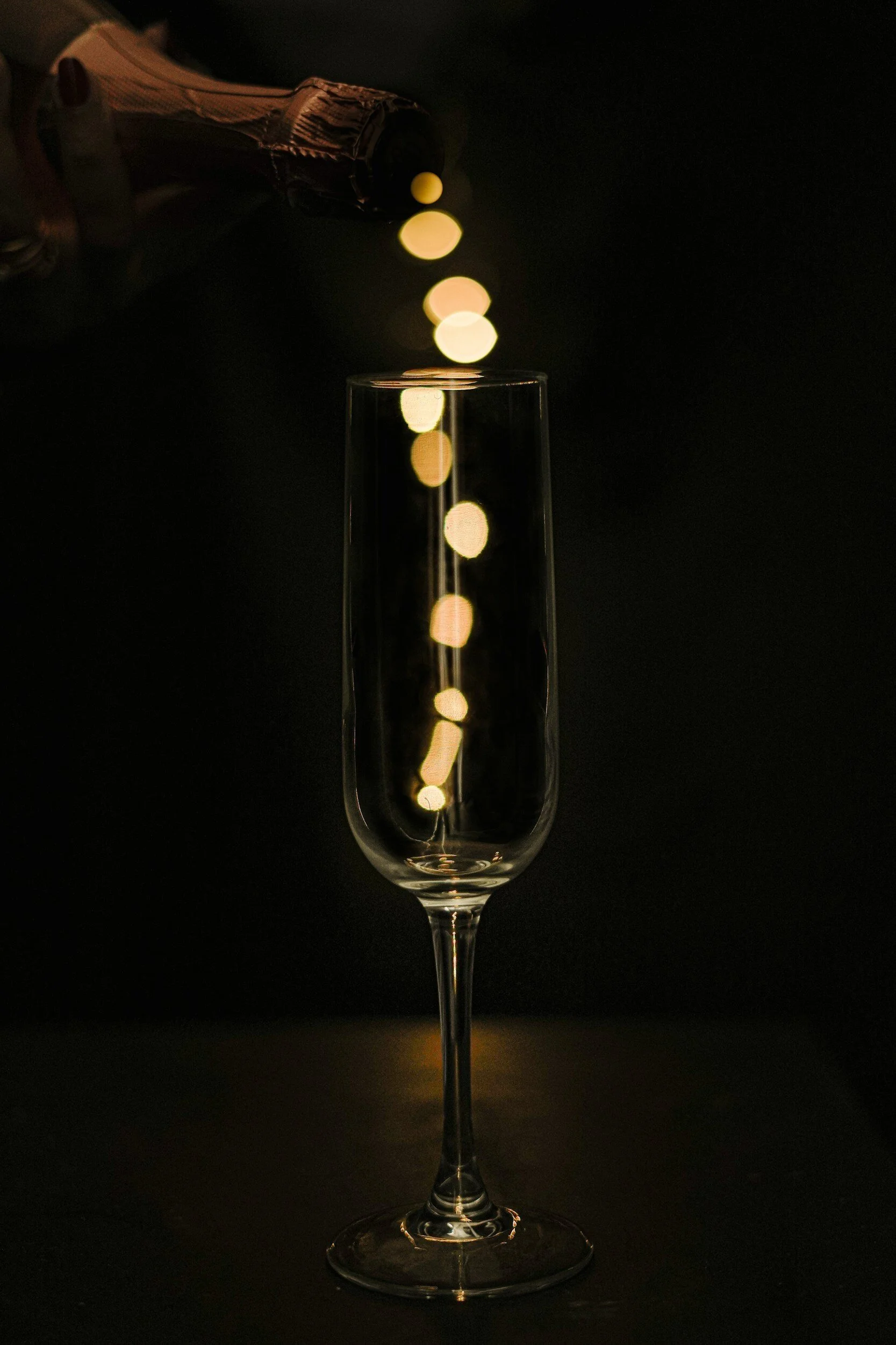 Champagne being poured into a tall, empty champagne flute with a dark background and bokeh lights reflected in the glass.