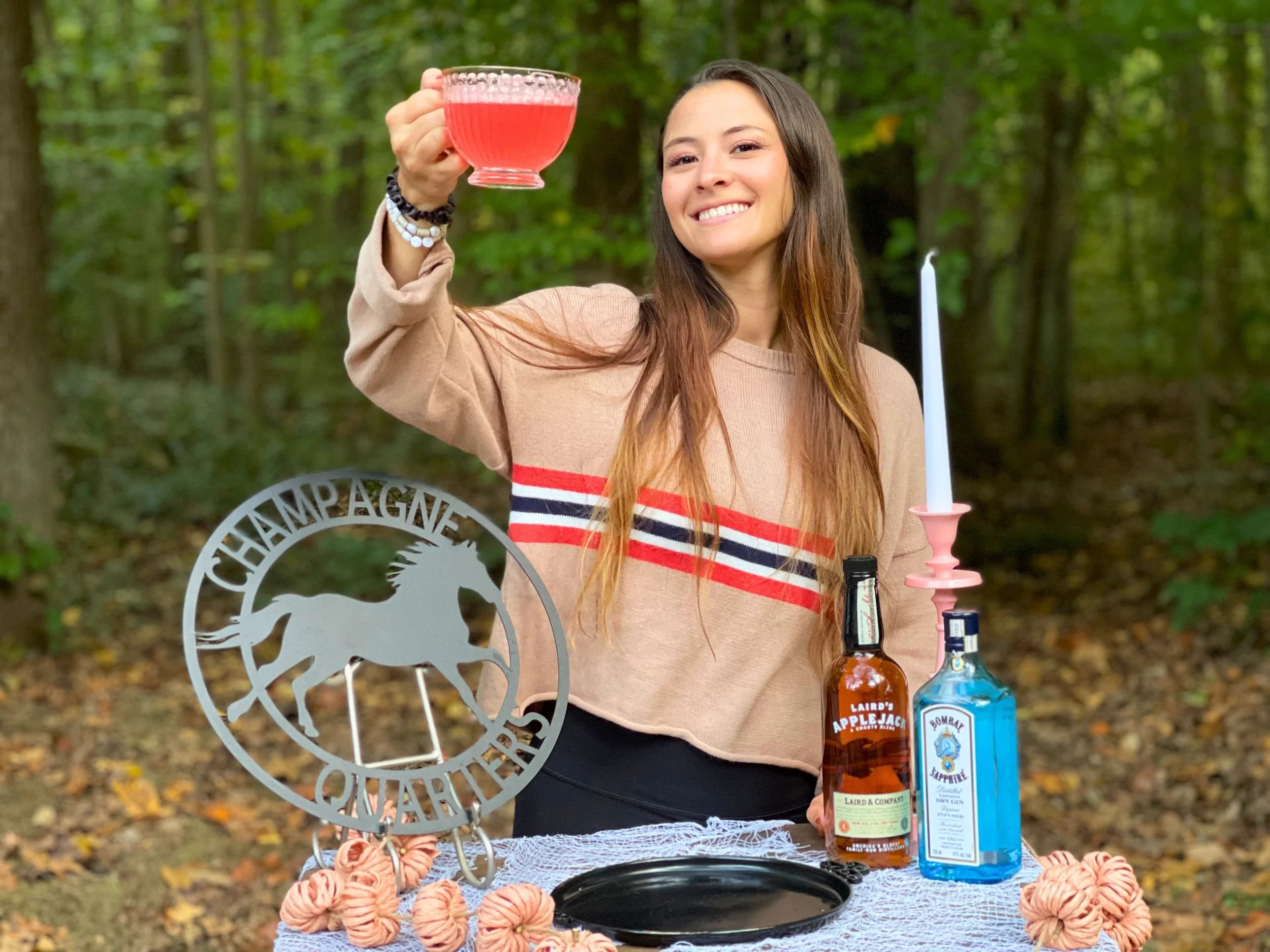 A young woman in a beige sweatshirt with red and black stripes is smiling and raising a glass of red drink in an outdoor forest setting. On the table in front of her are bottles of apple jack whiskey and Bombay Sapphire gin, a pink candlestick with a