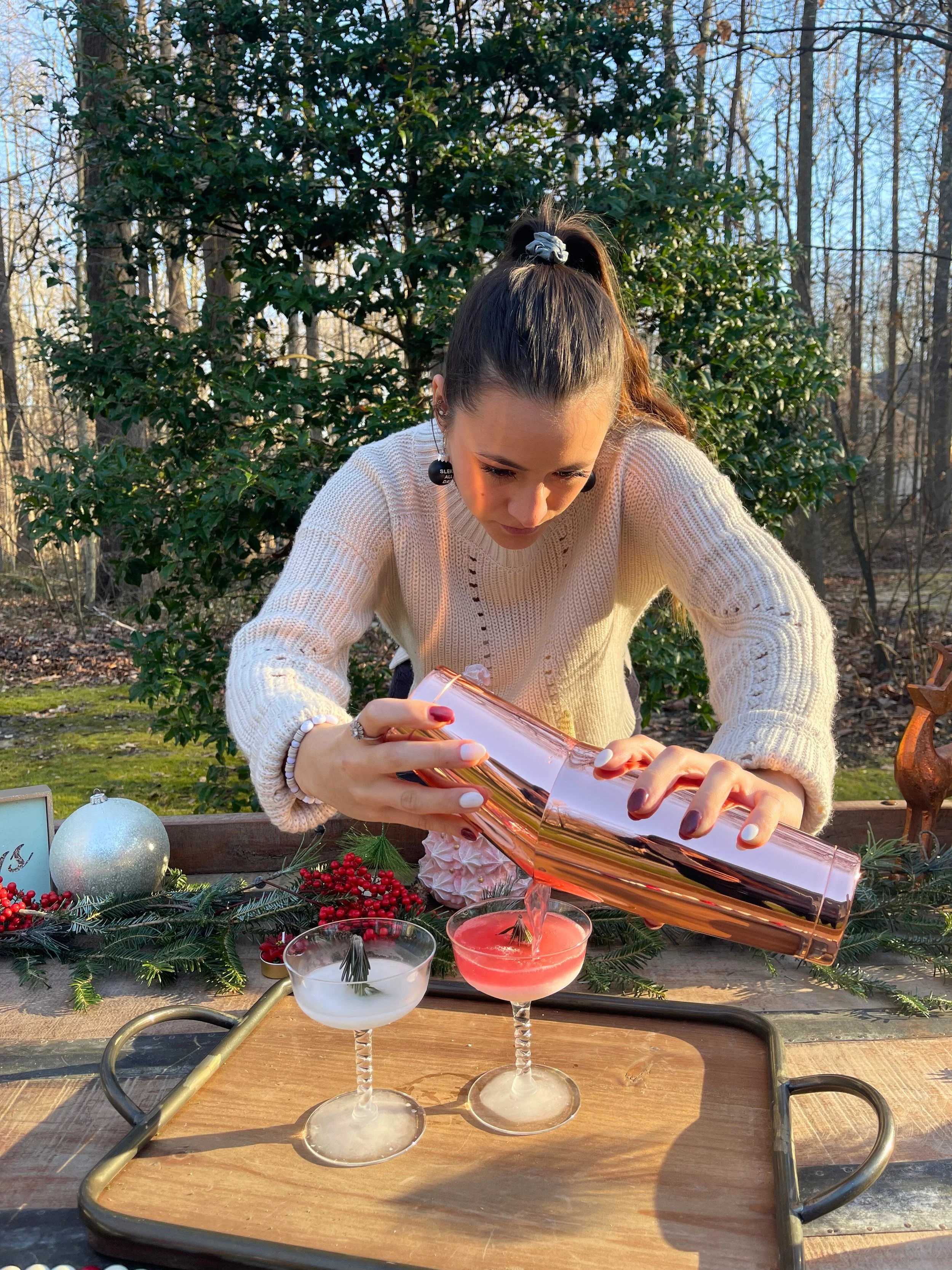 Woman in a white sweater pouring a pink cocktail into a glass outdoors decorated with holiday ornaments and greenery.