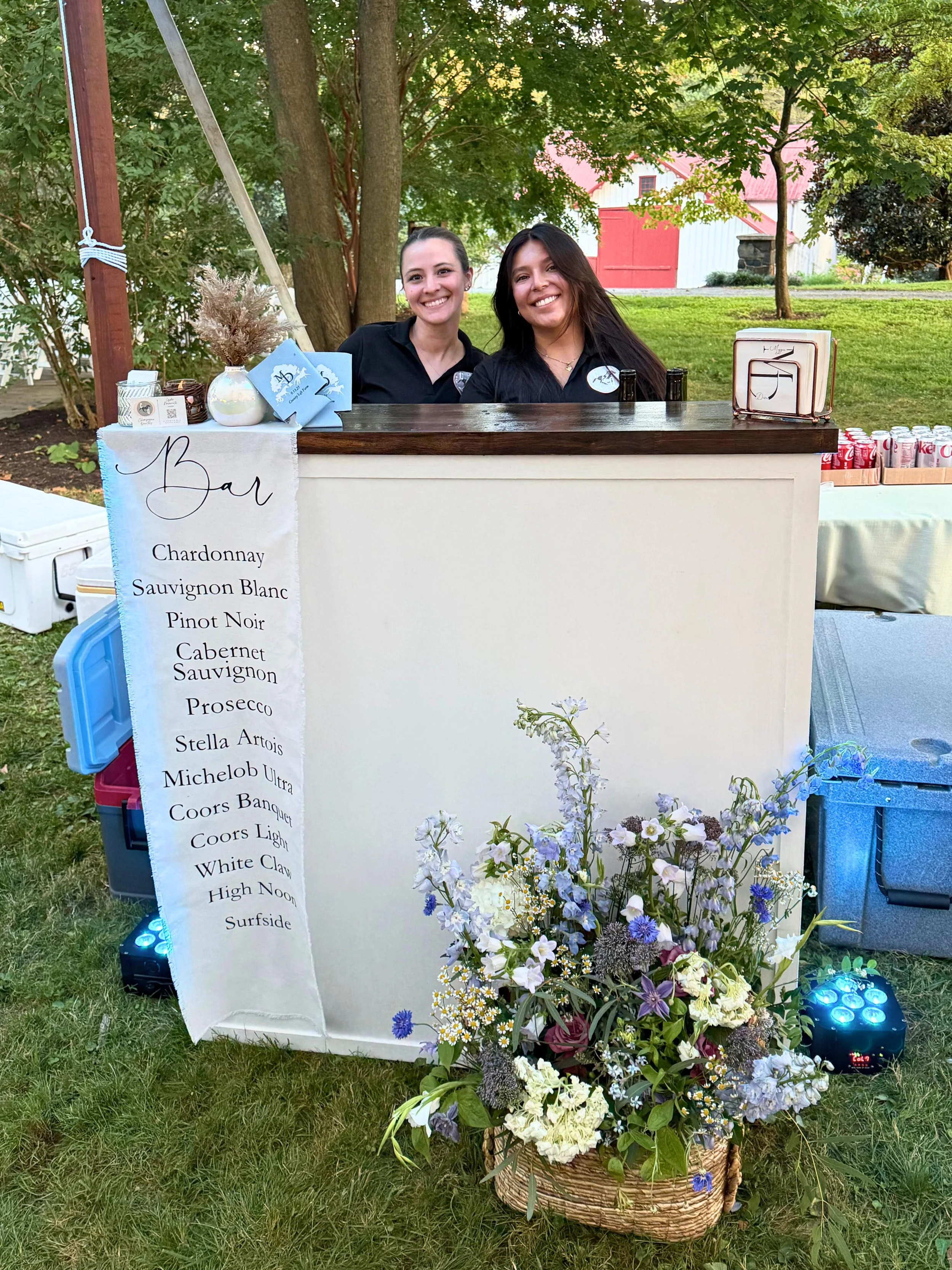 Two female bartenders standing behind a white wooden bar with an espresso butcher block counter. There is a fabric bar menu and a bouquet of flowers on the floor. This is one of the bar offerings from Champagne Quarters mobile bars for weddings, etc.