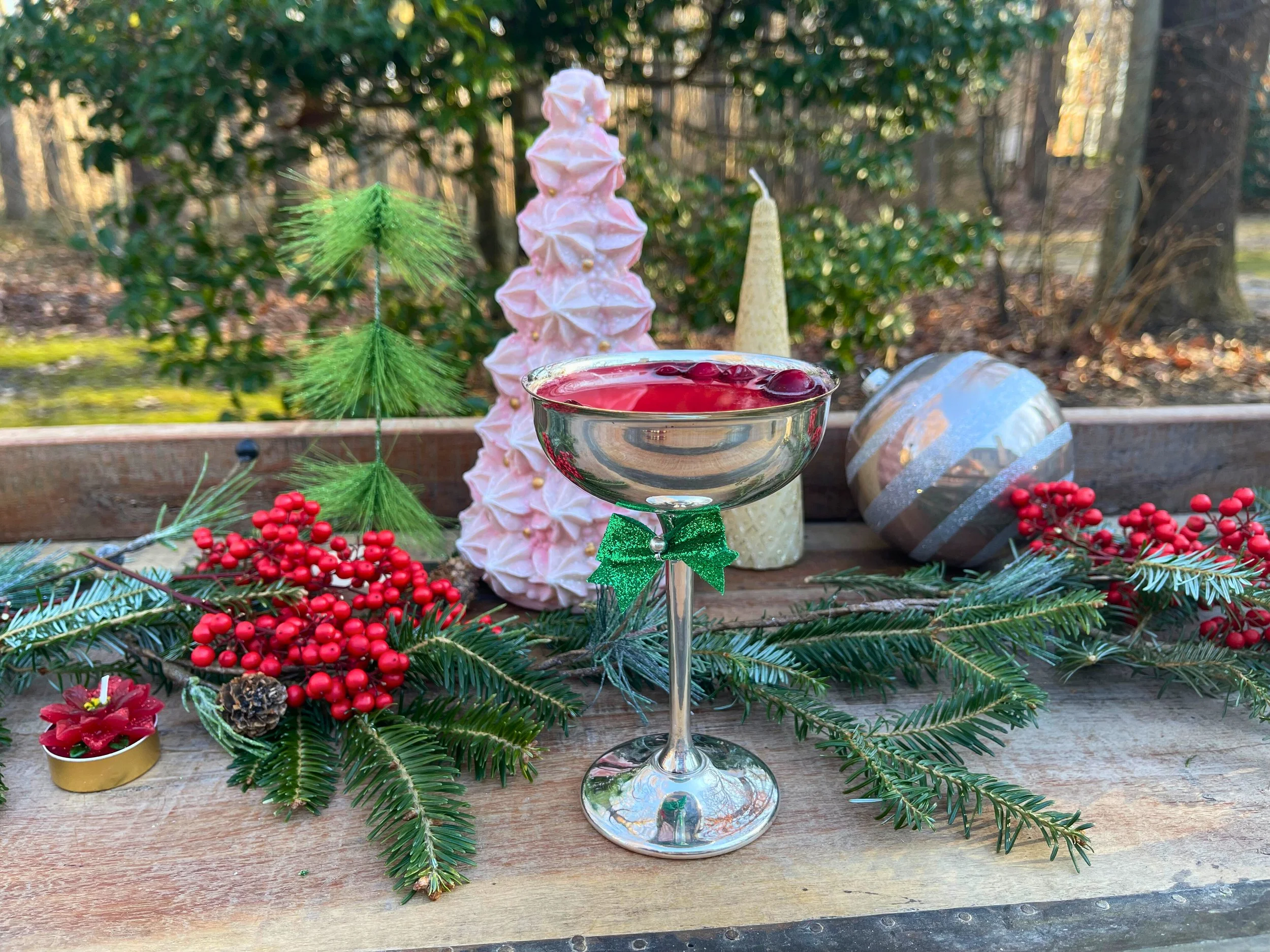 Christmas decorations including evergreen branches, red berries, pinecones, a pink ornament tree, a striped ornament, a white candle, and a glass cup of red punch on a wooden surface outdoors.
