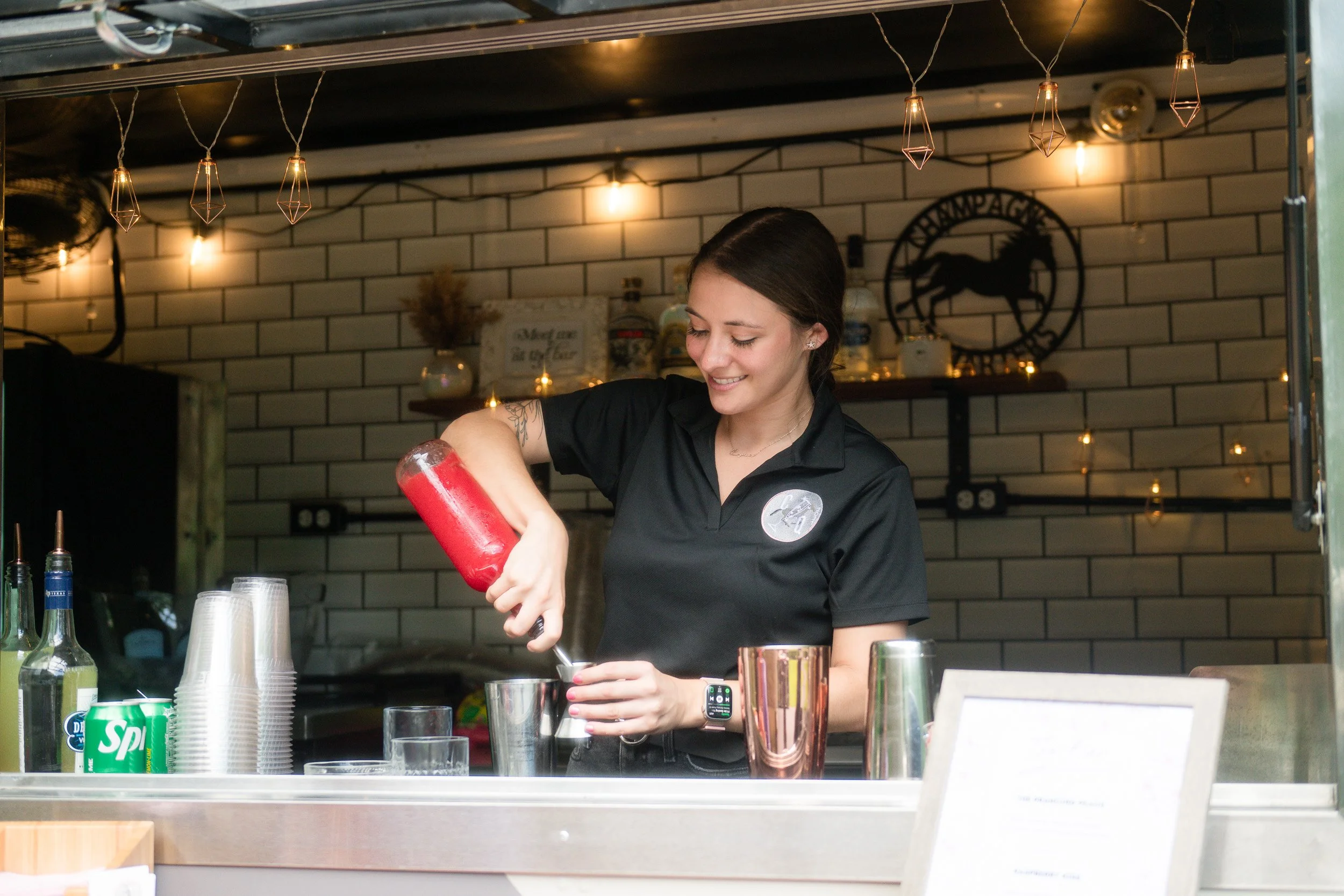A woman in a black shirt preparing a drink at a bar, pouring red syrup into a glass with bar tools and glasses on the counter, in a dimly lit setting with string lights overhead and a black horse and circle logo on the wall.