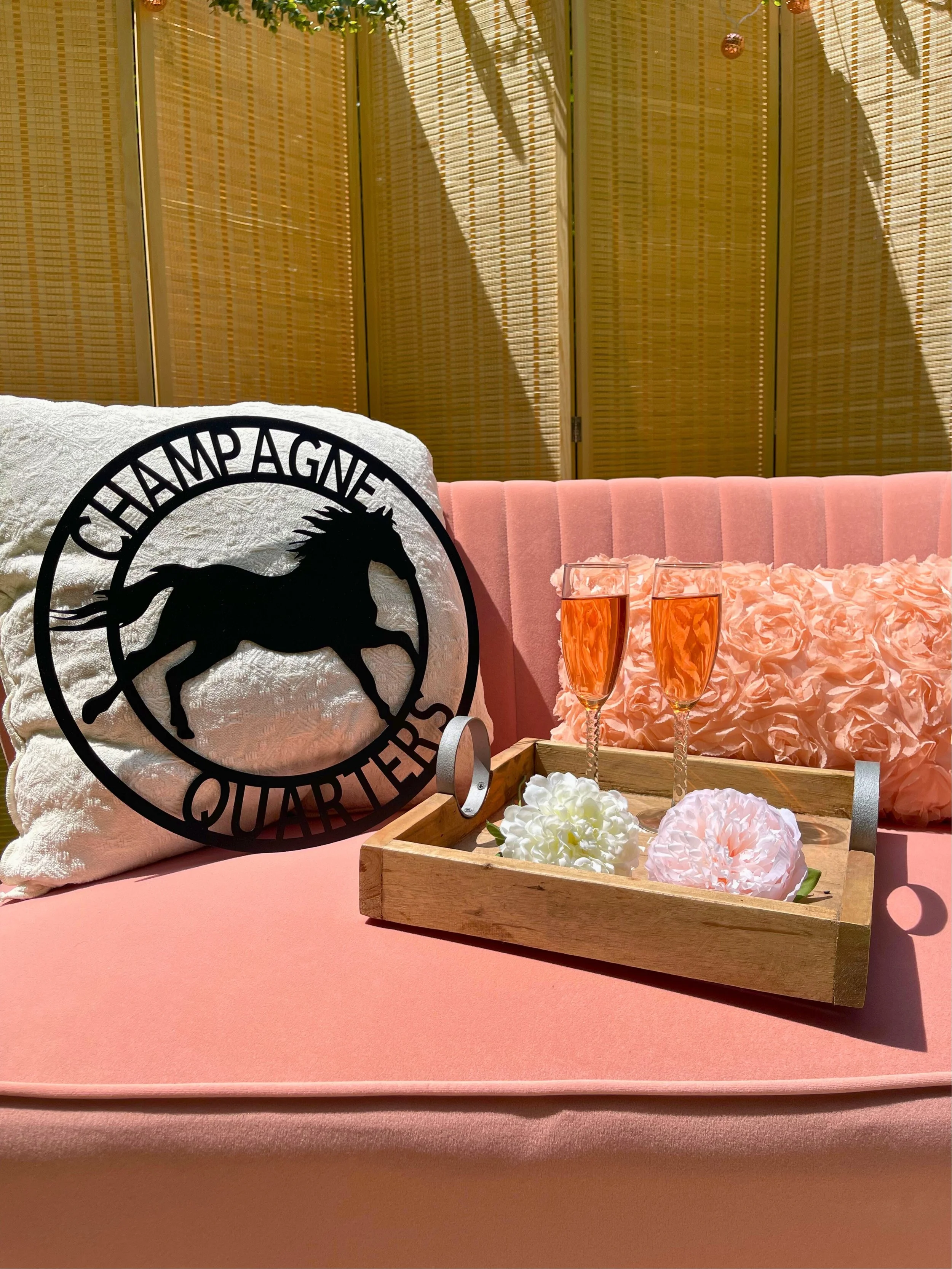 A pink sofa with a large white pillow featuring a black silhouette of a horse and the text ‘Champagne Quarters’. On the sofa, there is a pink, fluffy pillow and a wooden tray with two glasses of pink champagne, white and pink decorative flowers, and 