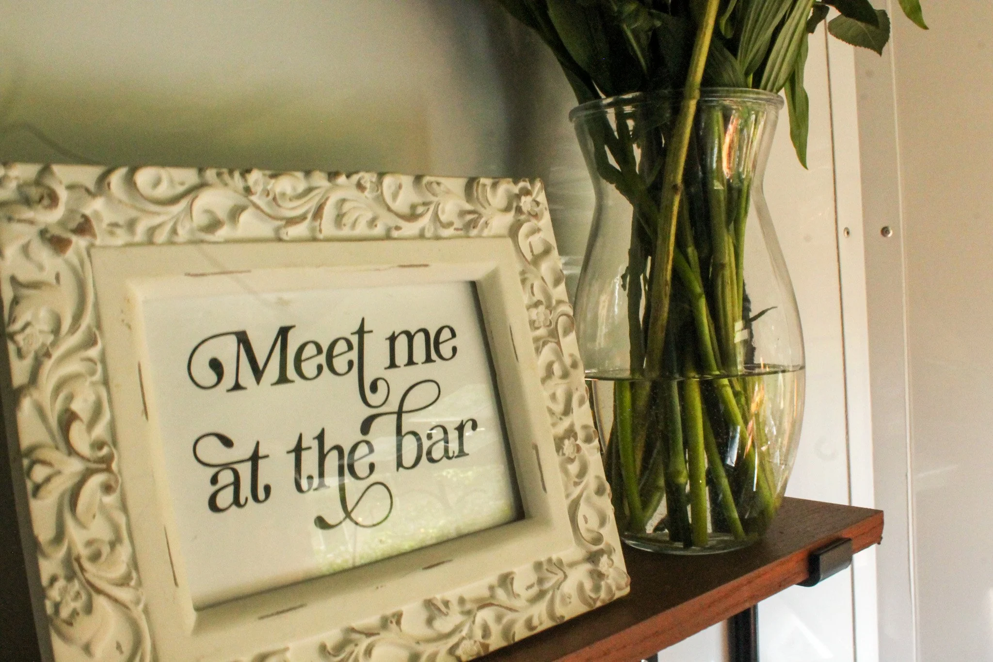 Decorative white frame with the message 'Meet me at the bar' written inside, placed on a wooden shelf beside a tall glass vase filled with water and green leafy stems.