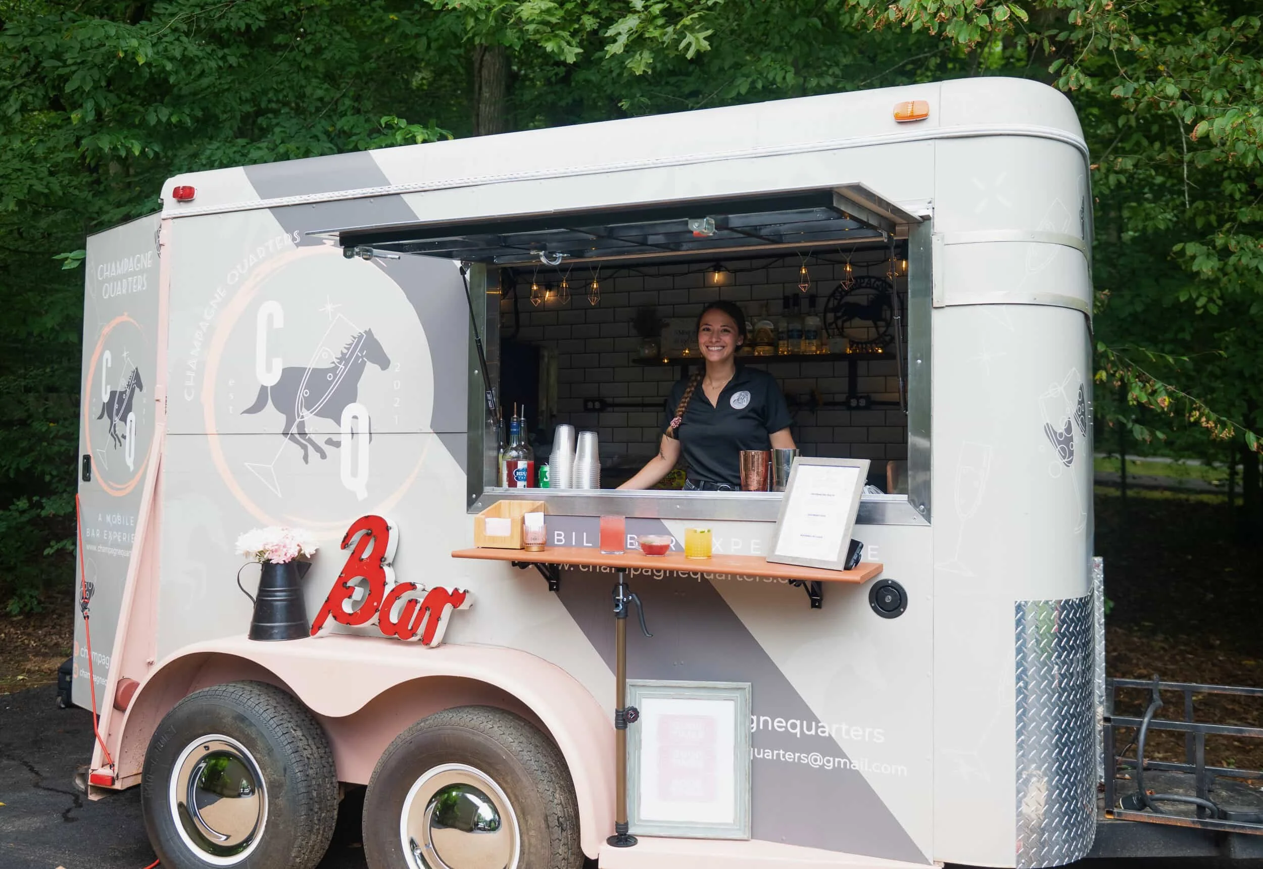 Female bartender standing in a mobile bar horse trailer, named Champagne Quarters, ready to serve drinks at a private event.