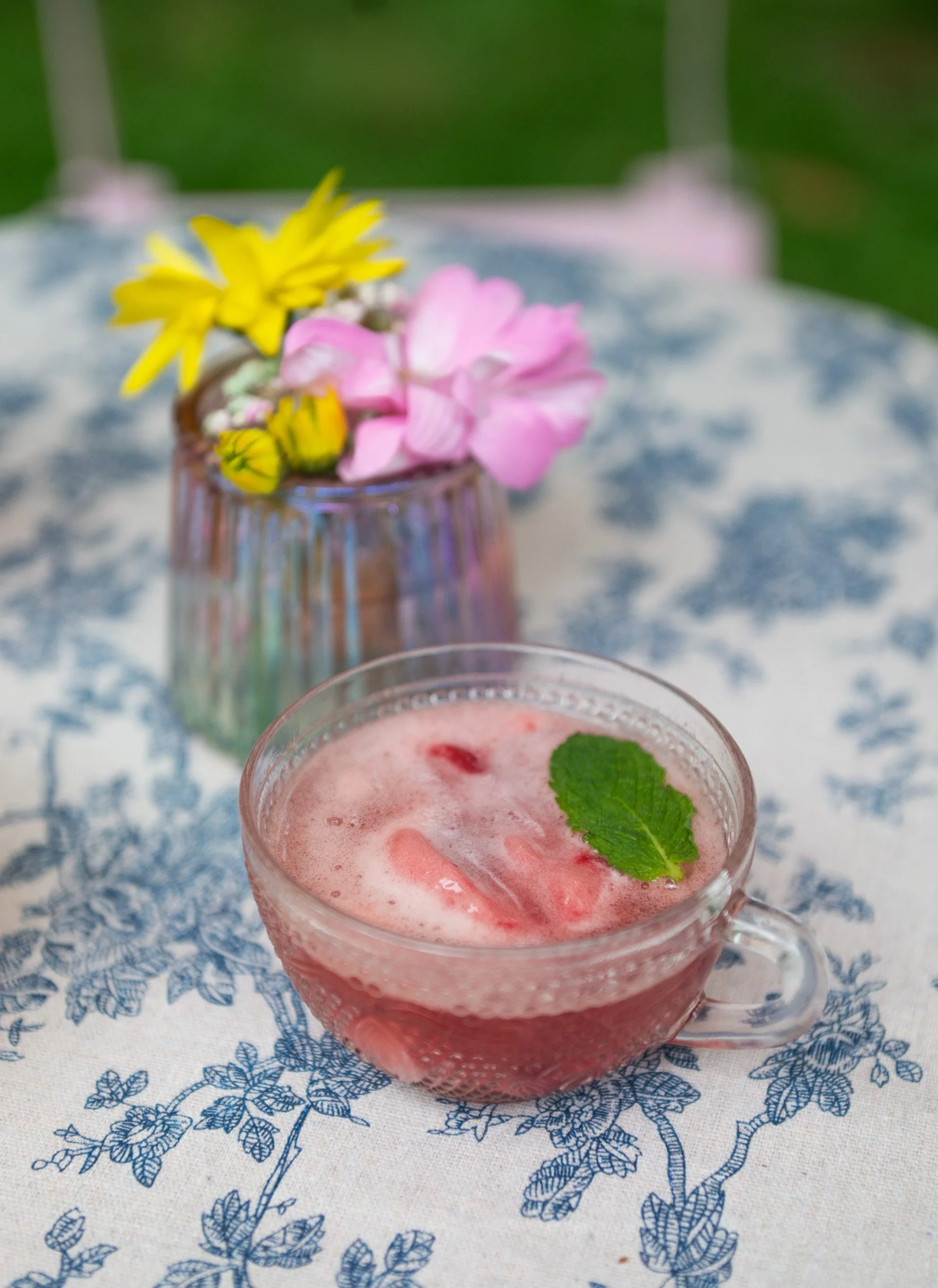 Glass cup of pink strawberry beverage garnished with a mint leaf on a tablecloth, with a small flower arrangement in the background.