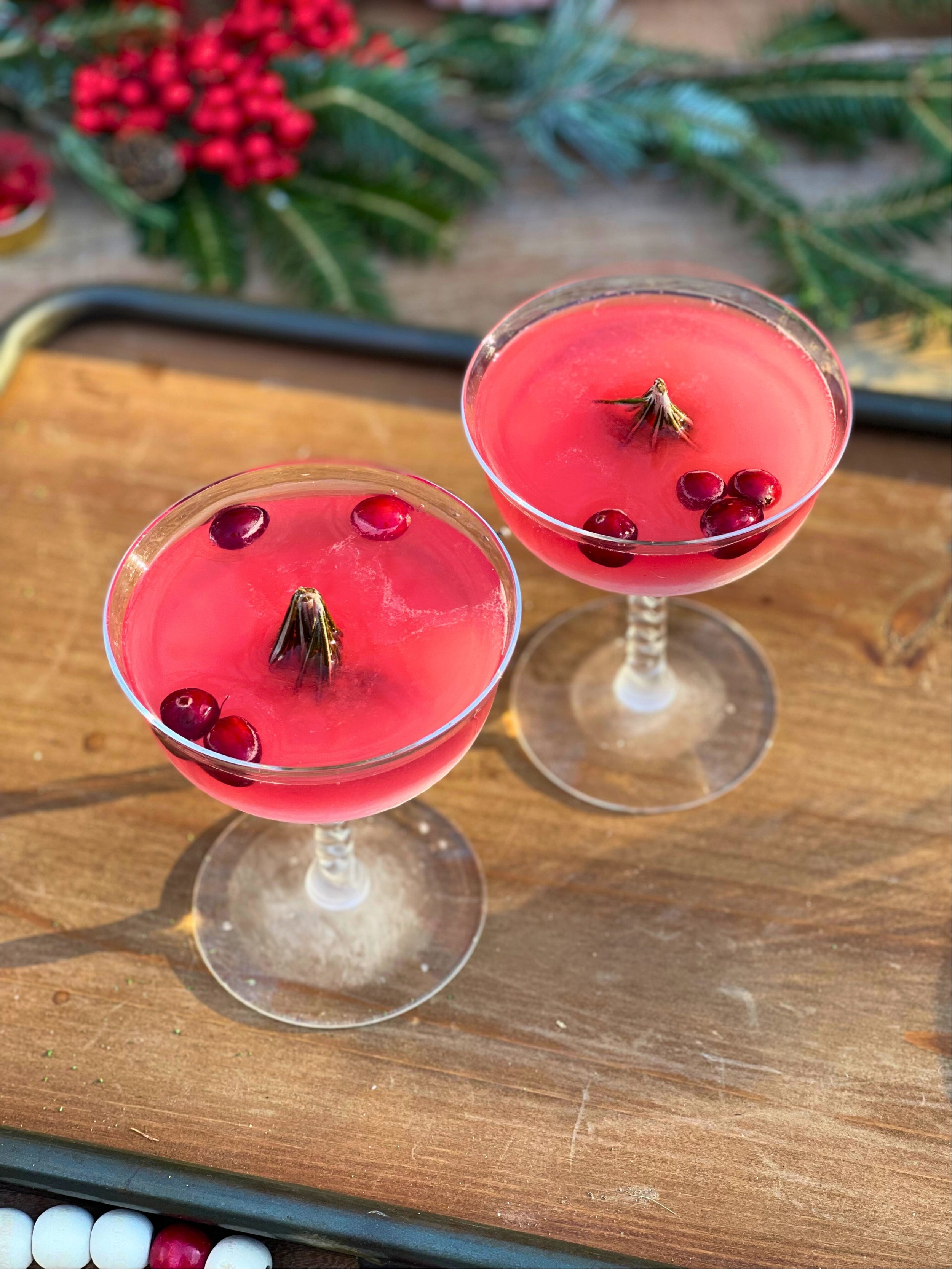 Two pink cocktails garnished with cranberries and a small pinecone in each glass, placed on a wooden surface with festive greenery in the background.