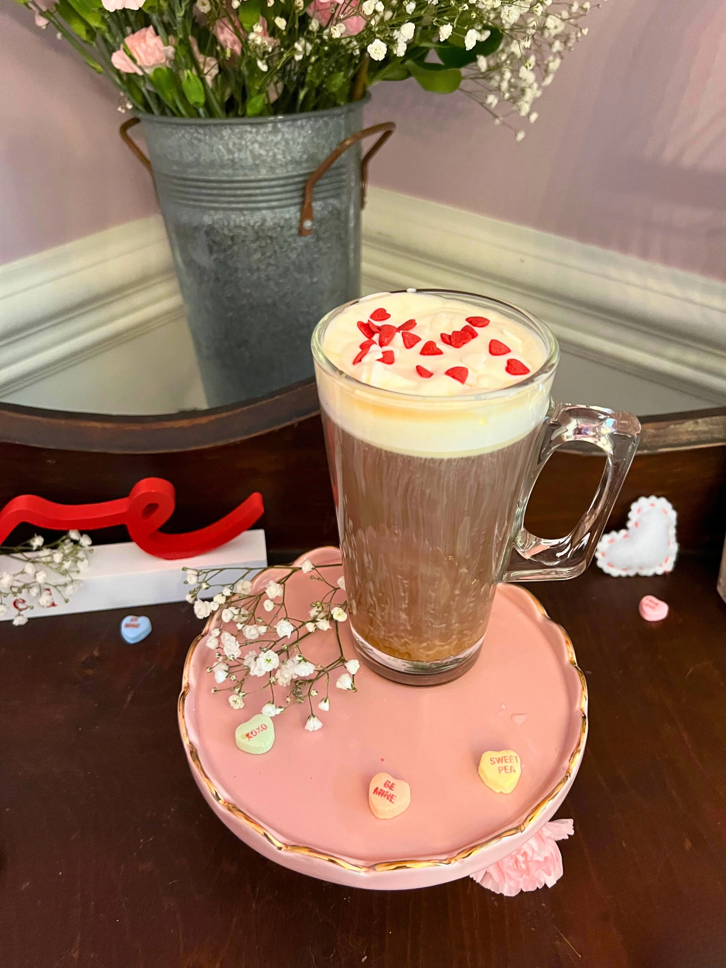 A glass mug of hot chocolate topped with whipped cream and red heart-shaped sprinkles, placed on a pink, decorated ceramic plate with small Valentine's Day messages and white baby's breath flowers. In the background, a vase of pink and white flowers 