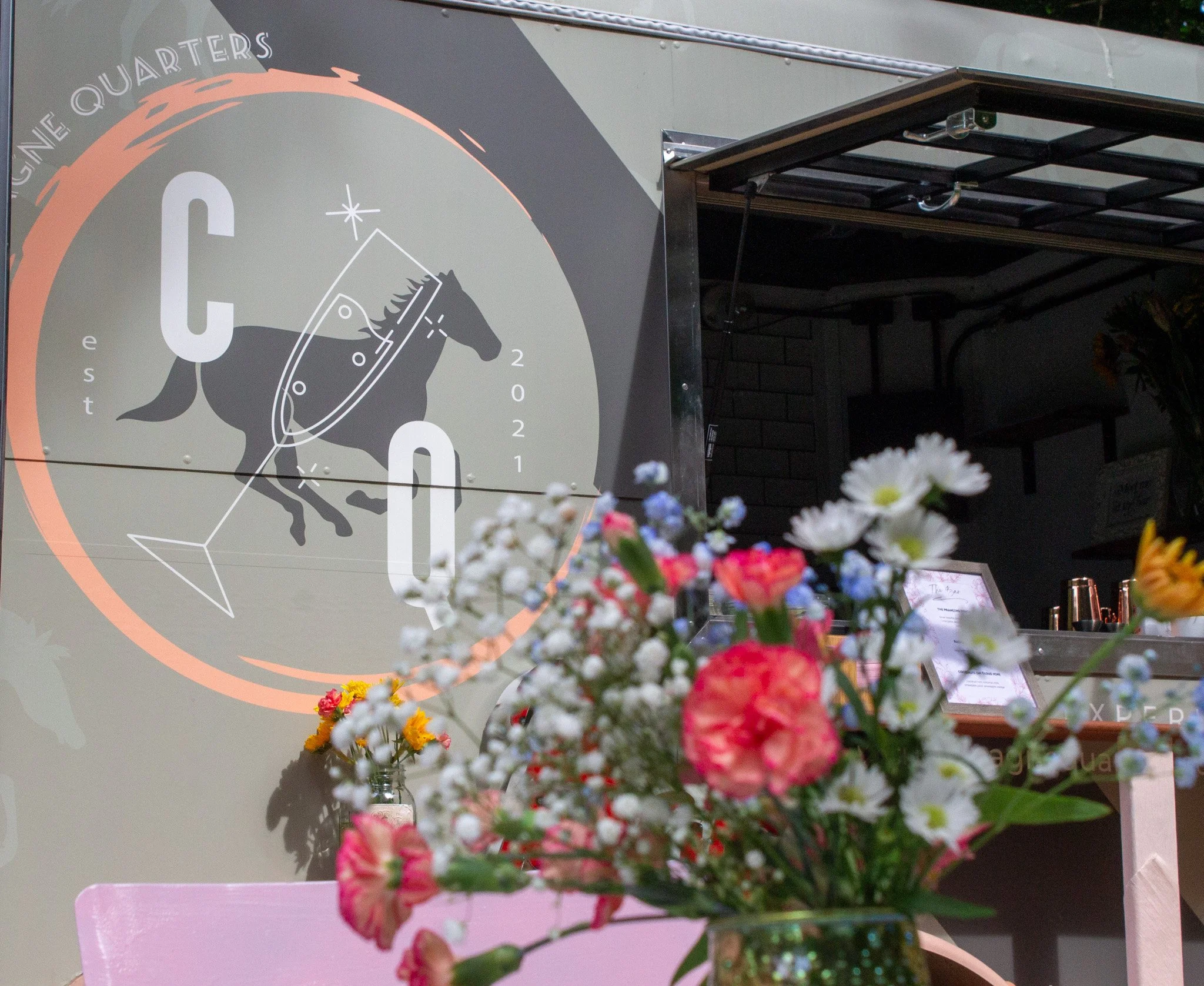 A sign with a horse silhouette, text reading "CHAMPAGNE QUARTERS," and the year 2021, with flowers in the foreground.