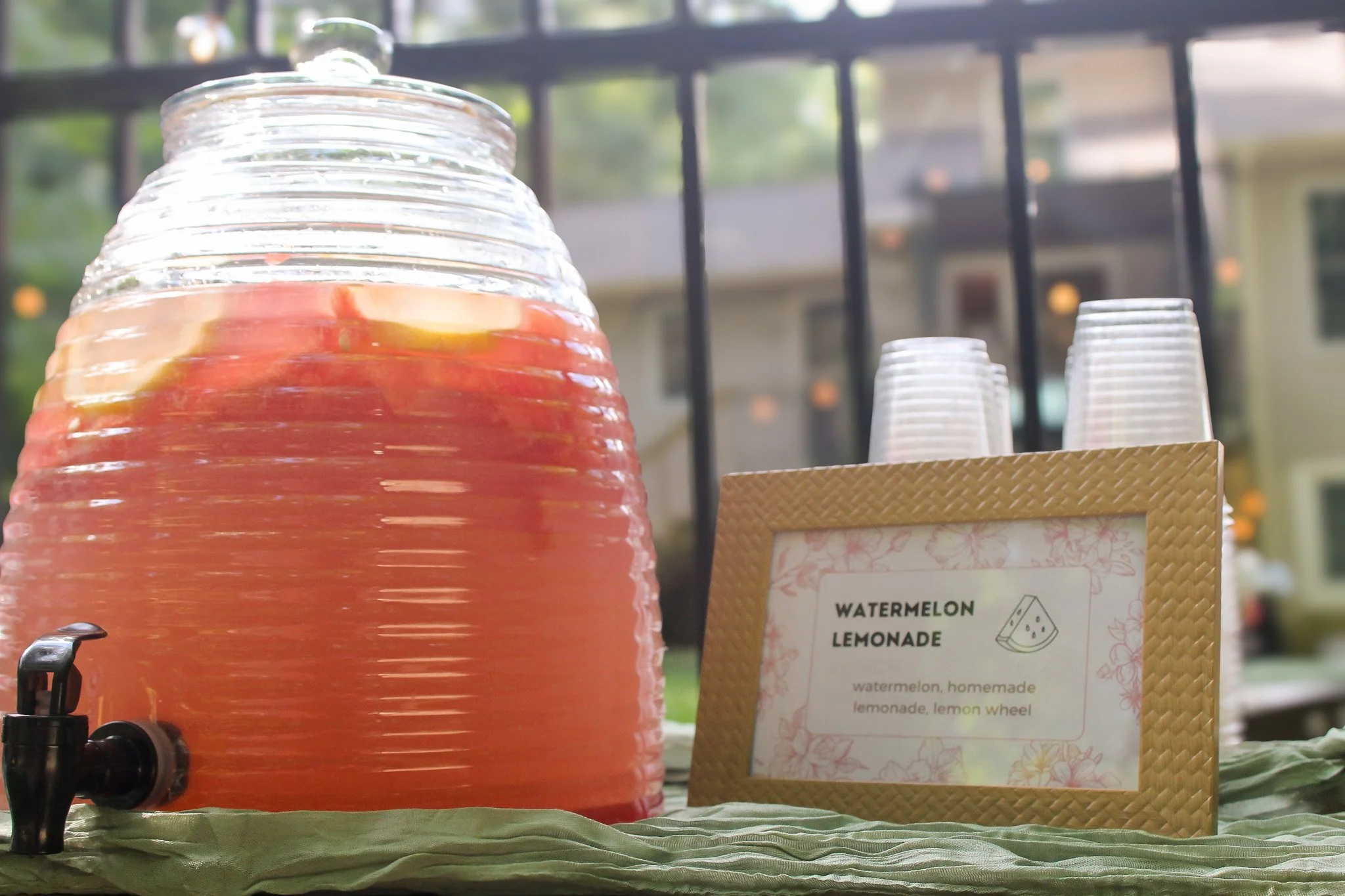 Large glass dispenser filled with pink watermelon lemonade, with a small black spout, on a green tablecloth outdoors. Next to it, clear plastic cups and a sign that reads 'Watermelon Lemonade' describing the drink.