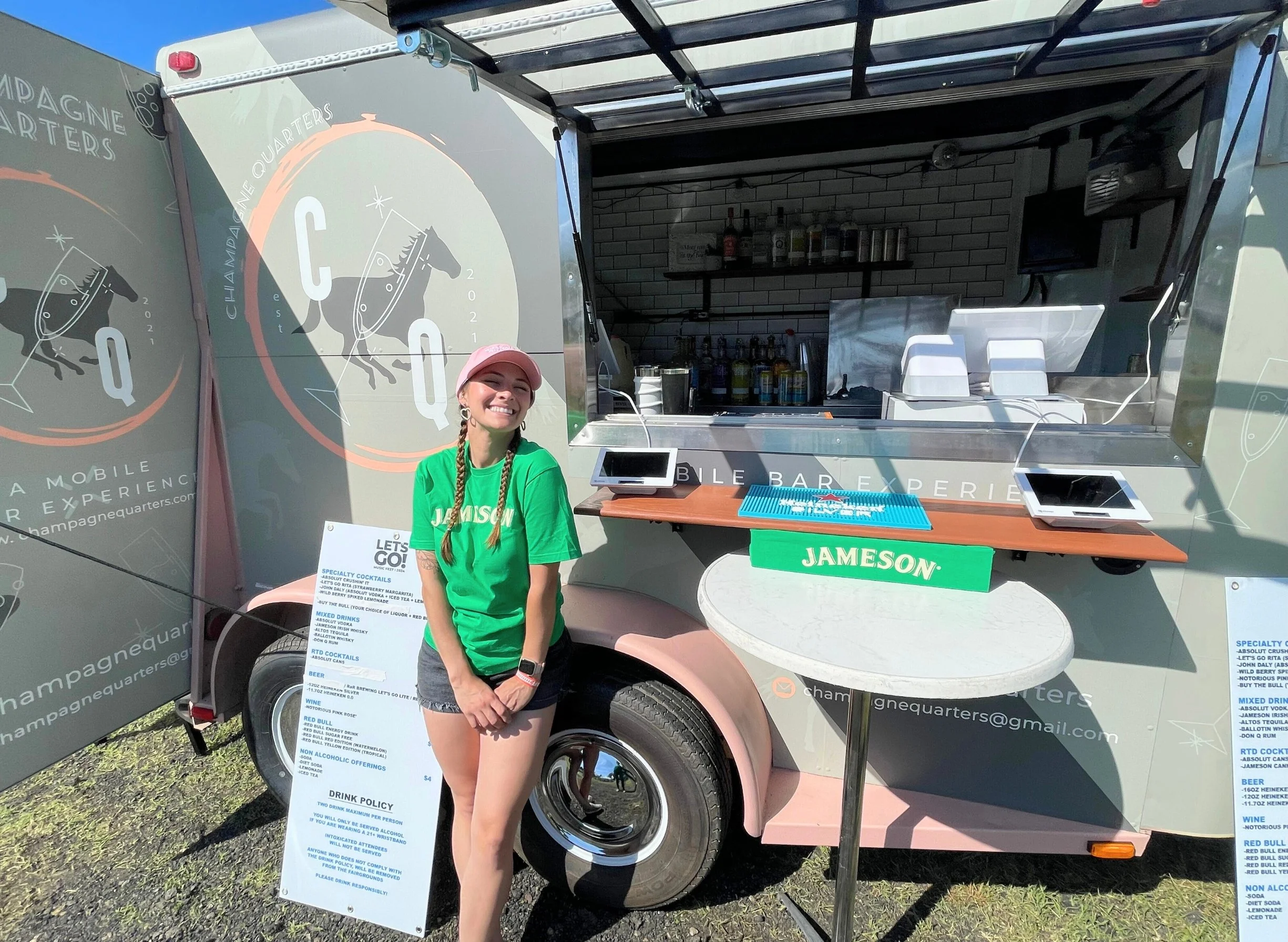 A woman in a green T-shirt and pink cap standing in front of a mobile bar trailer with a menu sign and a Jameson logo, smiling and enjoying the outdoor setting.