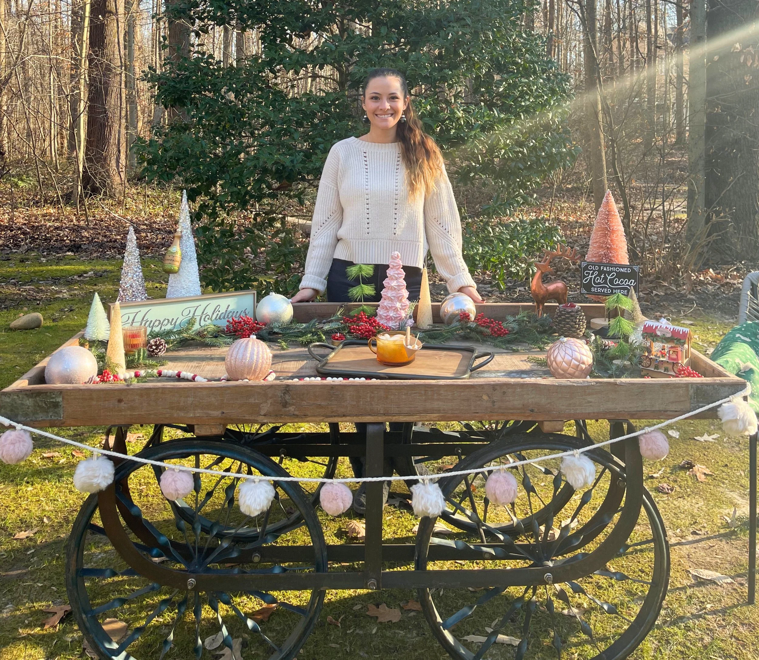 Woman standing behind a decorated wooden cart outdoors with holiday decorations including ornaments, trees, and a sign that says 'Happy Holidays' and 'Old Fashioned Hot Cocoa Served Here', in a wooded area with sunlight filtering through trees.