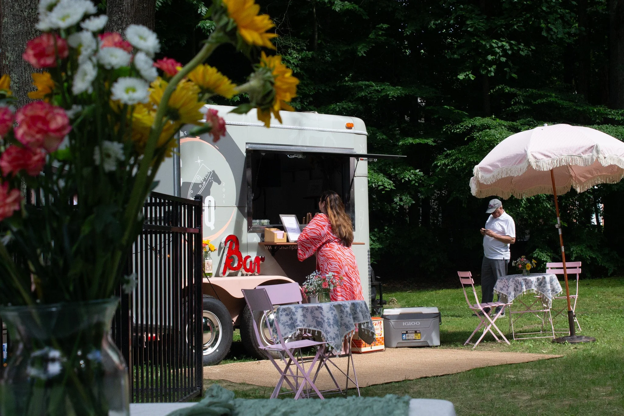 A food truck with a woman ordering at the counter, standing outdoors with tables and chairs under a pink umbrella, and a man standing nearby looking at his phone. There are trees in the background.