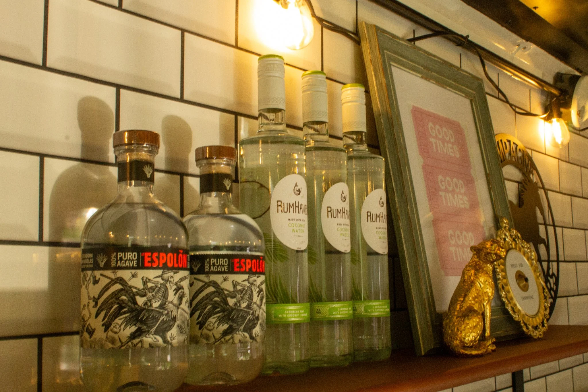 Bottles of alcohol, including Espolon tequila and RumHaven coconut water, on a wooden shelf against a white tiled wall in a bar or restaurant setting, with framed decor and warm lighting.