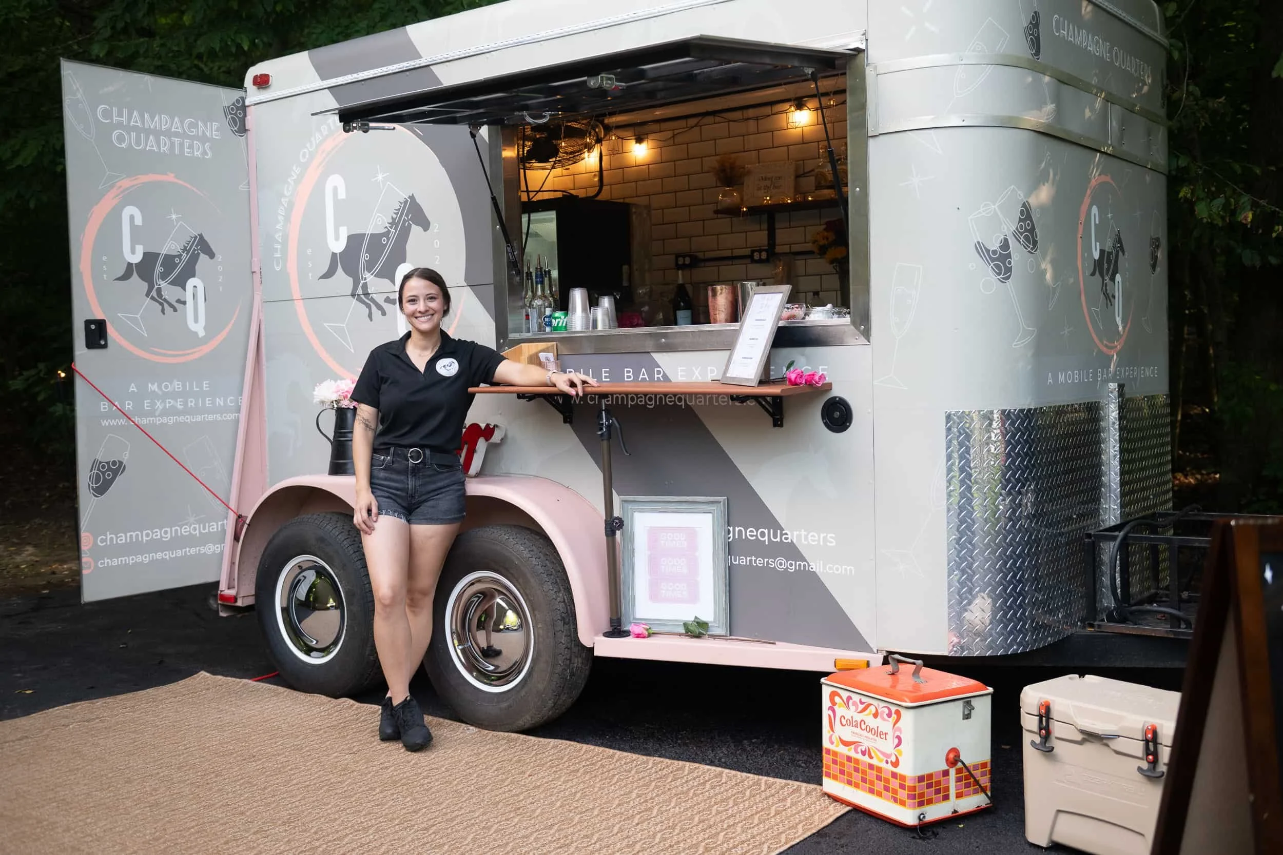 Female bartender standing in front of horse trailer mobile bar, named Champagne Quarters, a mobile bar experience serving Maryland and Virginia.