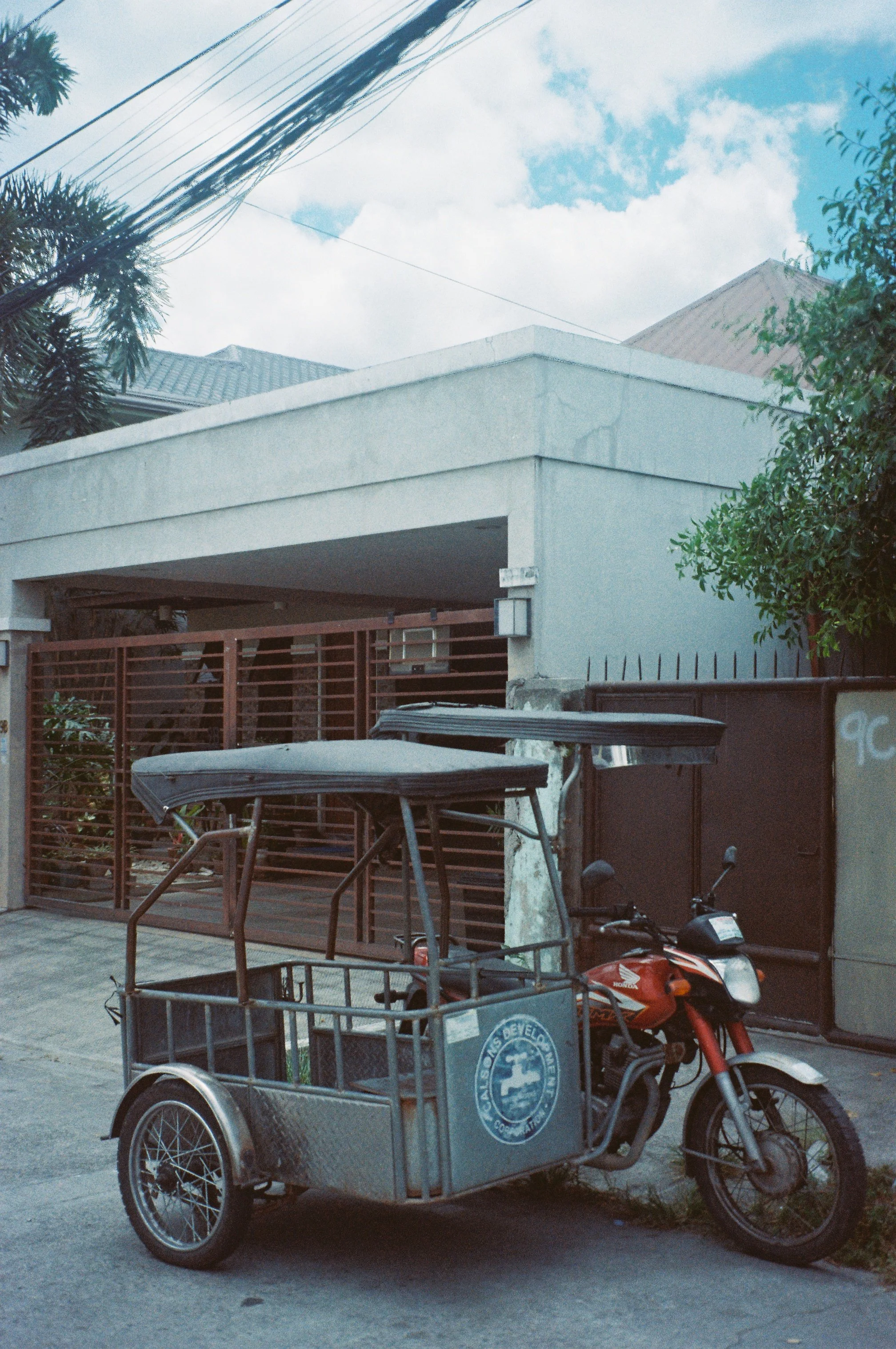 A three-wheeled cargo bike with a large front compartment, parked on a street in front of a house with a garage. The house has a white exterior, a grey roof, and a red gate. A motorcycle is parked beside the bike. Overhead, there are power lines and 