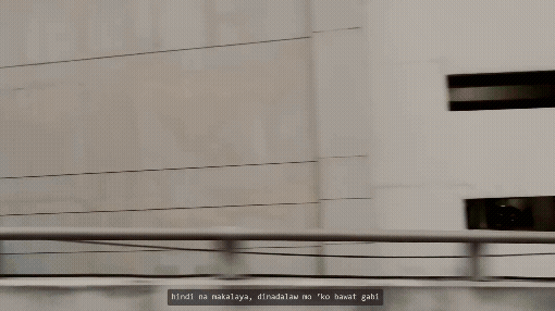 A view of a concrete building with horizontal slats and a railing in the foreground, with a subtitle in a foreign language at the bottom.