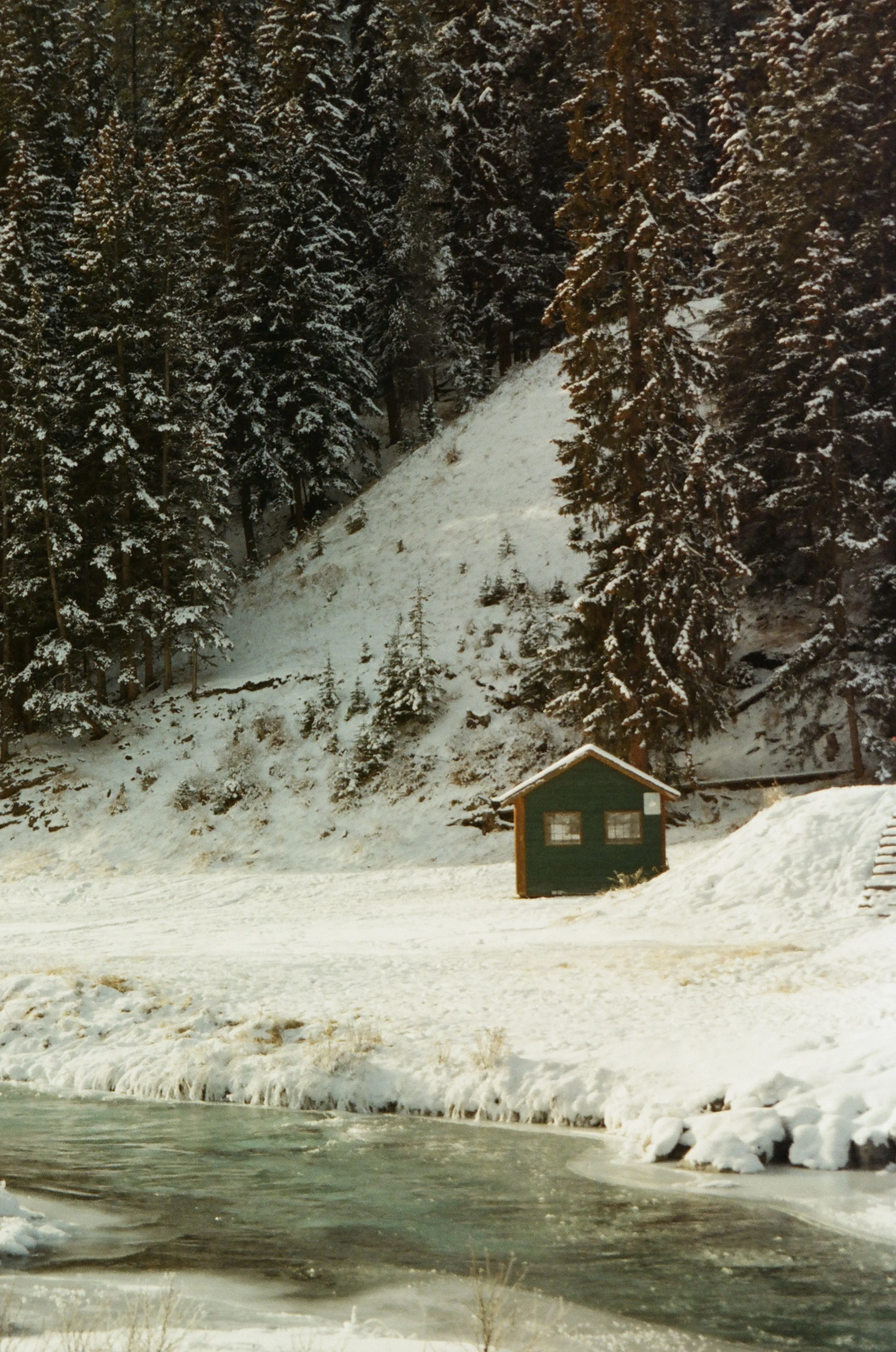 Snowy landscape with pine trees on a hillside, small green wooden house near a frozen river, and snow-covered ground.