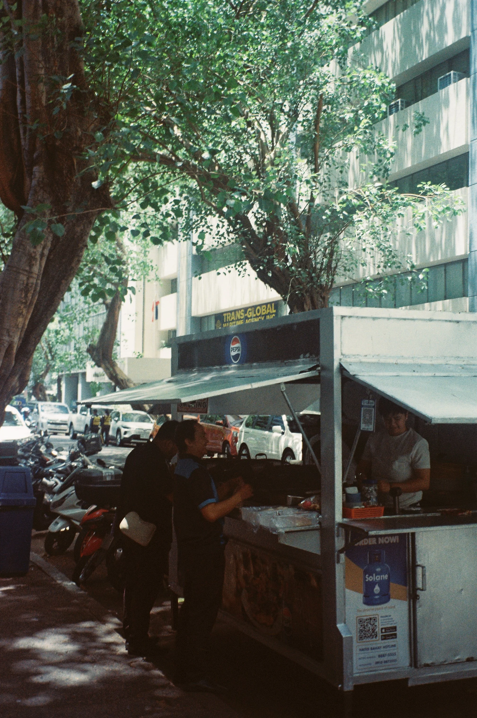 Street scene with a food cart, people eating and talking, parked motorbikes, cars, green trees, and a modern multi-story building in the background.