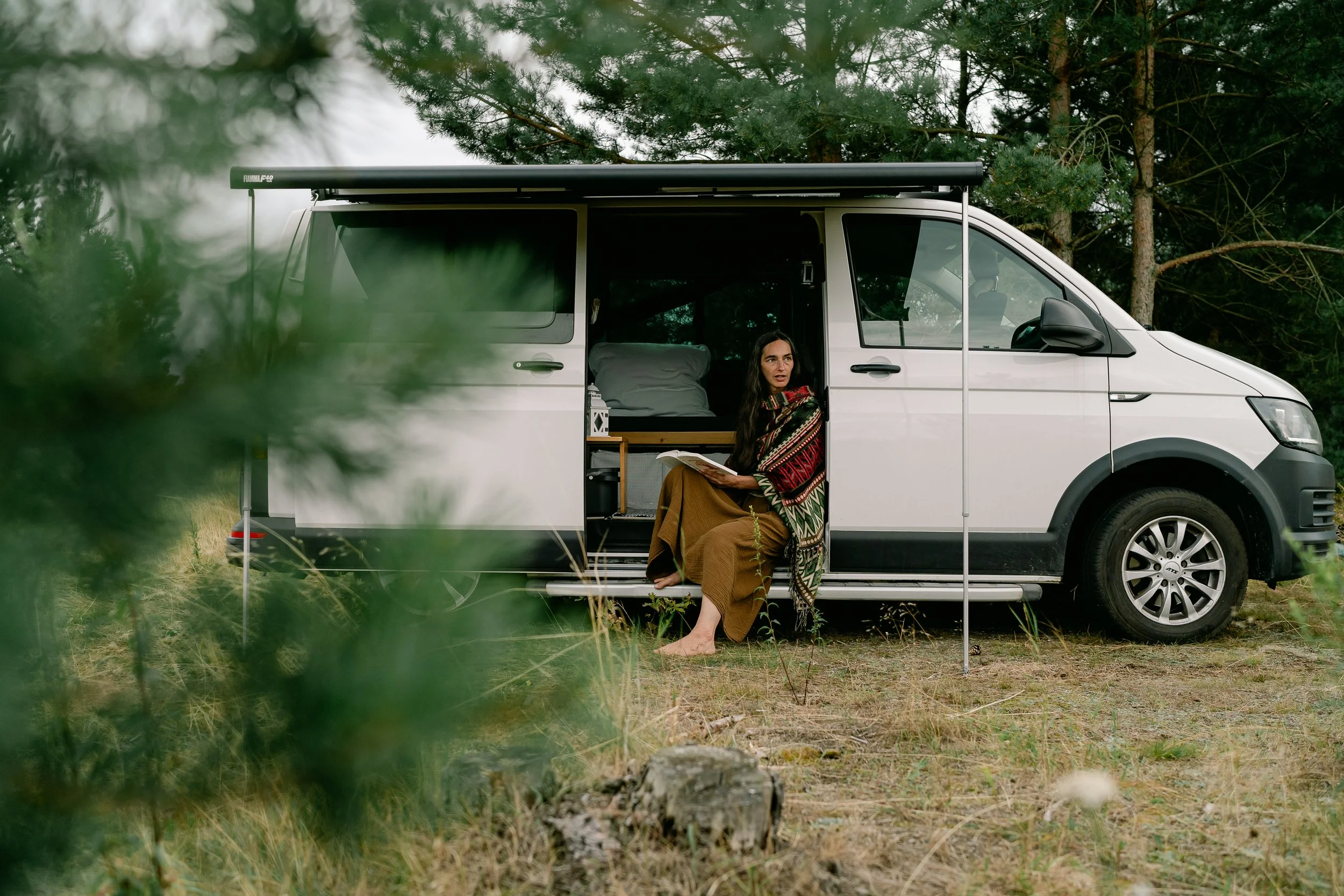 Woman reading in the open door of a campervan among pine trees, natural brand photography