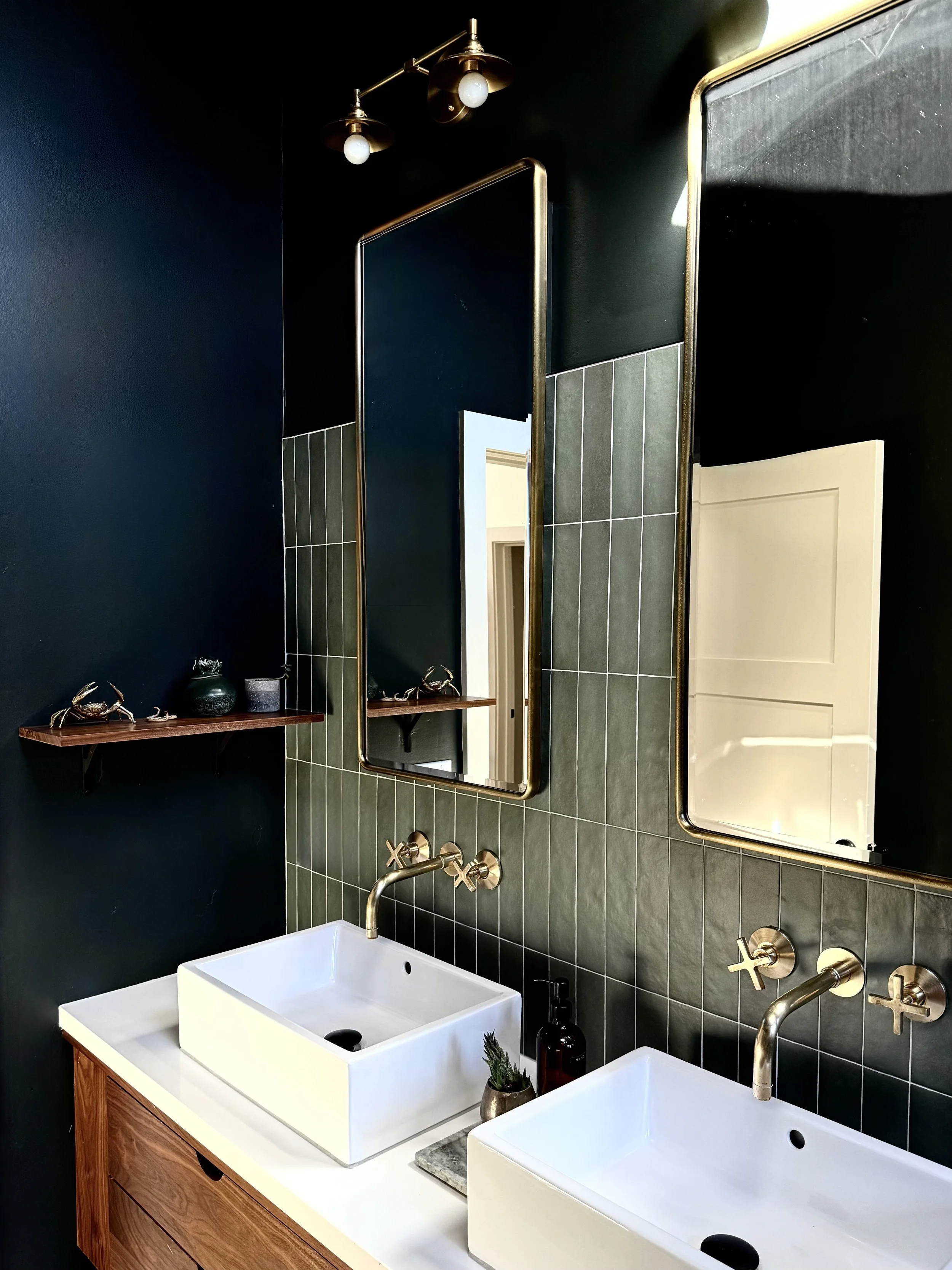 Modern bathroom with dark green walls, two white rectangular vessel sinks, gold wall-mounted faucets, two tall mirrors with gold frames, small wooden shelf with decor, and dark green tiles behind the sinks.