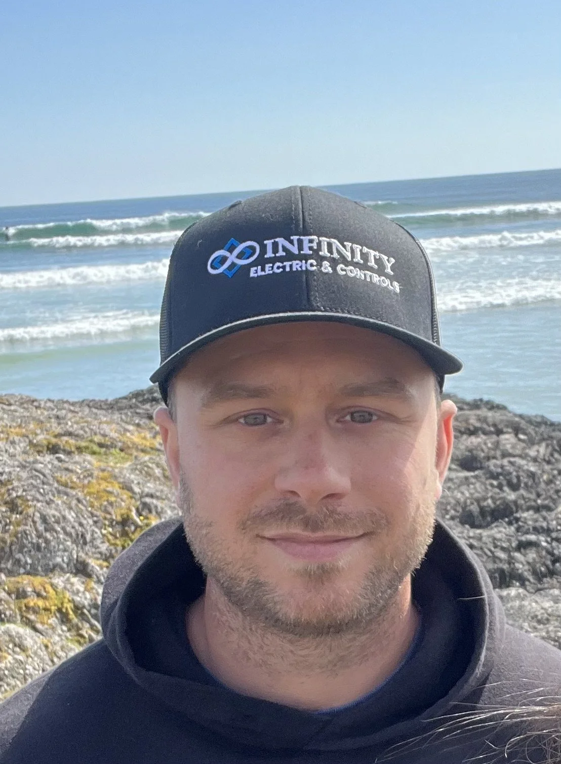 A man is taking a selfie on a rocky beach with the ocean and waves in the background. He is wearing a black cap with the logo and text 'INFINITY ELECTRIC & CONTROLS' and a black hoodie, in courtenay, bc. IESBC - infinity electrical services