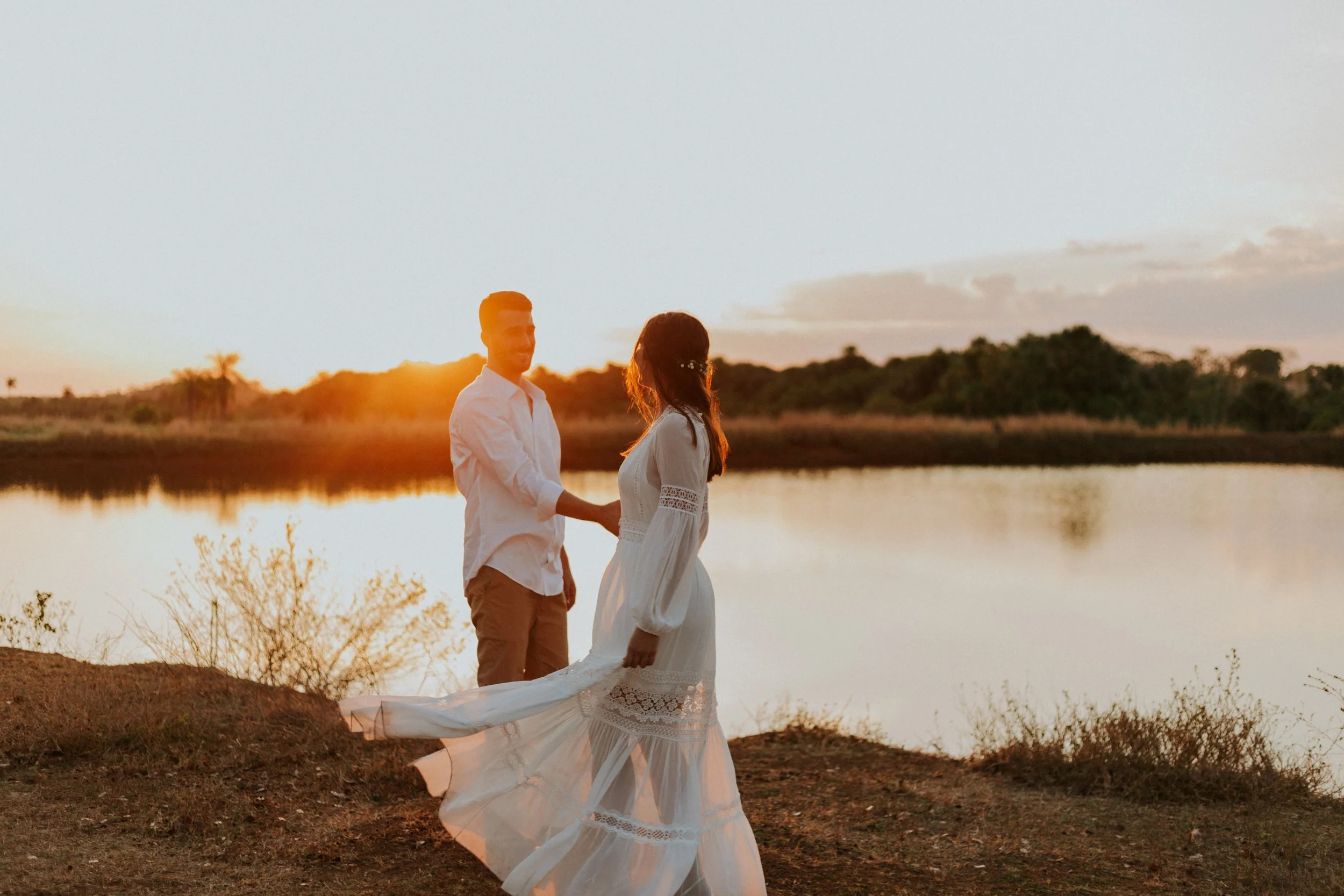 A couple holding hands by a lake at sunset, woman in a white flowing dress and man in a white shirt, romantic scene at Timeless Tree Studios, the best photography studio in Toronto.