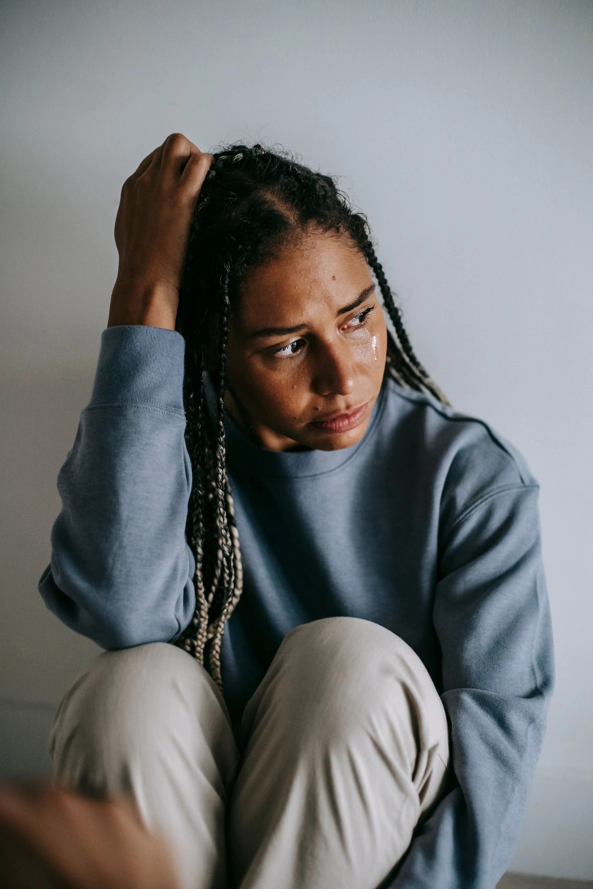 A young woman with curly dark hair and light brown skin sits against a plain light gray wall, gazing thoughtfully to the right, wearing a casual light blue sweatshirt and beige pants, with her right hand resting on her head.