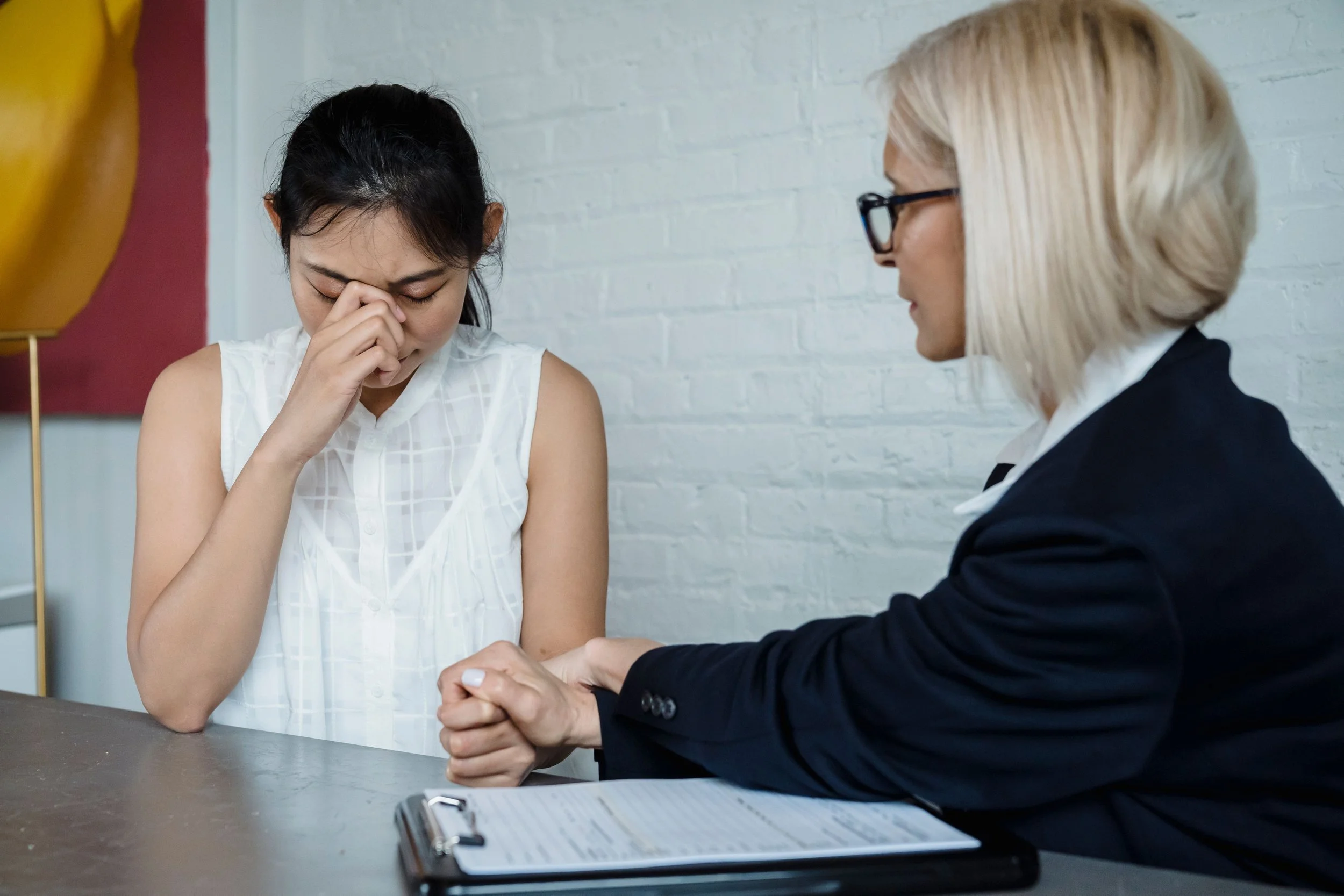 A woman with dark hair and a white sleeveless blouse appears distressed, holding her face with her eyes closed. Another woman with blonde hair, glasses, and a dark blazer is sitting next to her, holding her hand in a comforting manner in an indoor setting with a white brick wall in the background.
