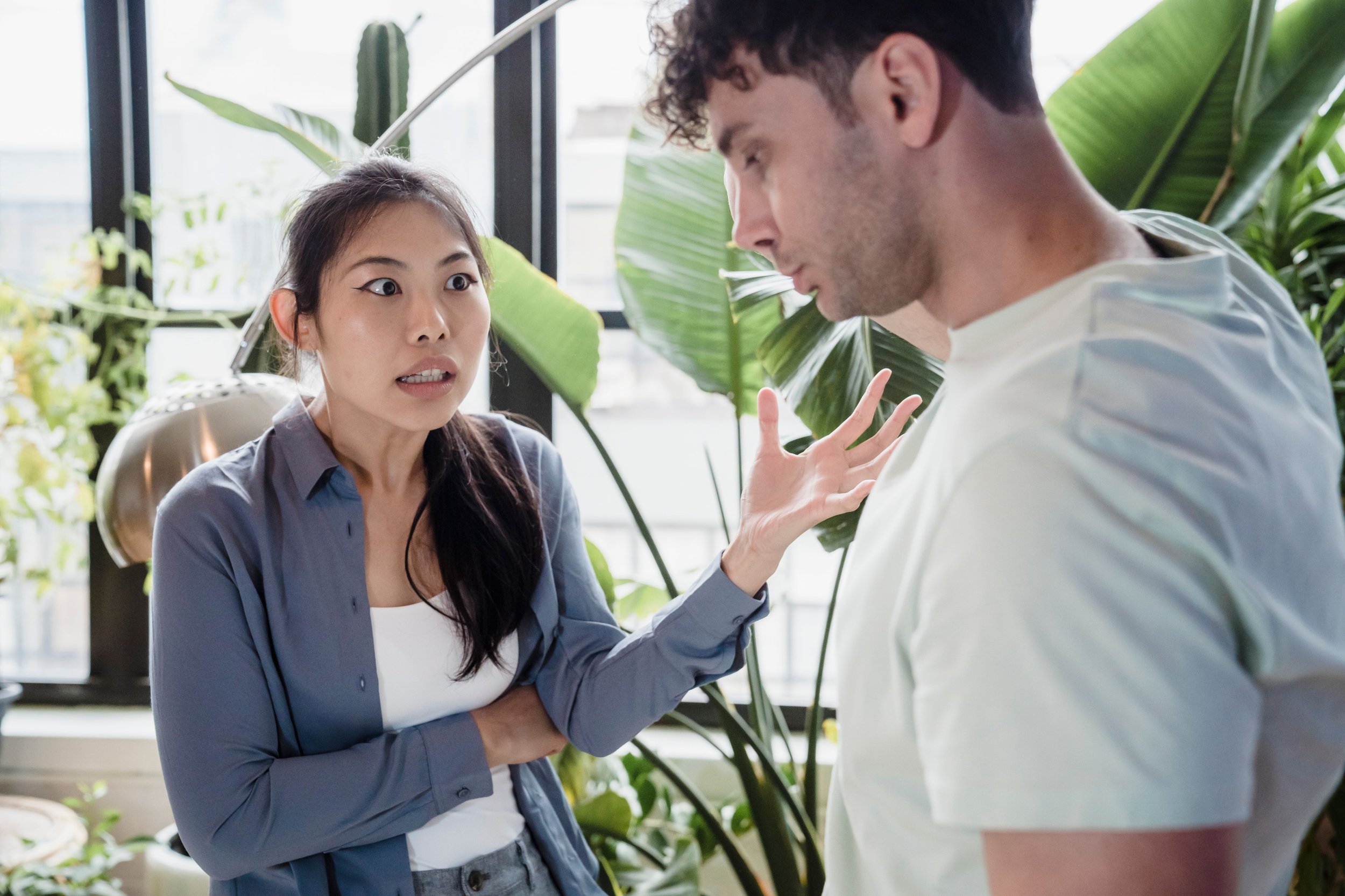 A woman with long dark hair and winged eyeliner appears to be arguing or speaking emphatically to a man with short curly hair, in a setting with large green plants and natural light.