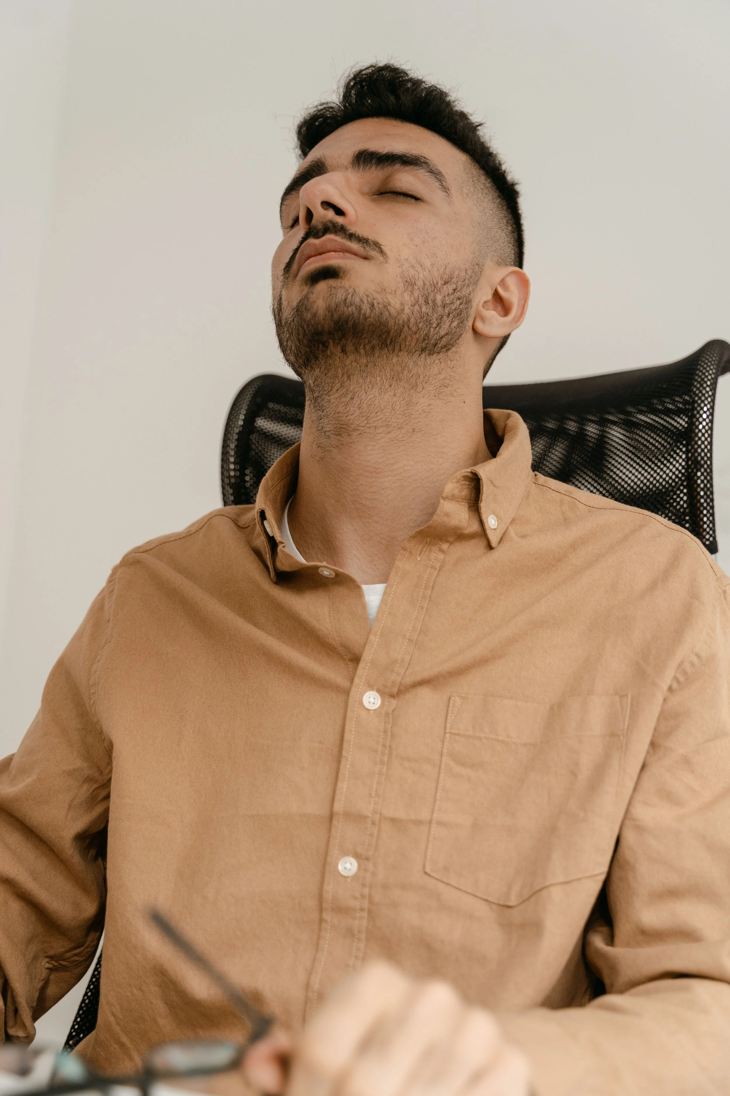 Young man with dark hair and beard, wearing a tan button-up shirt, sitting in an office chair with his eyes closed.