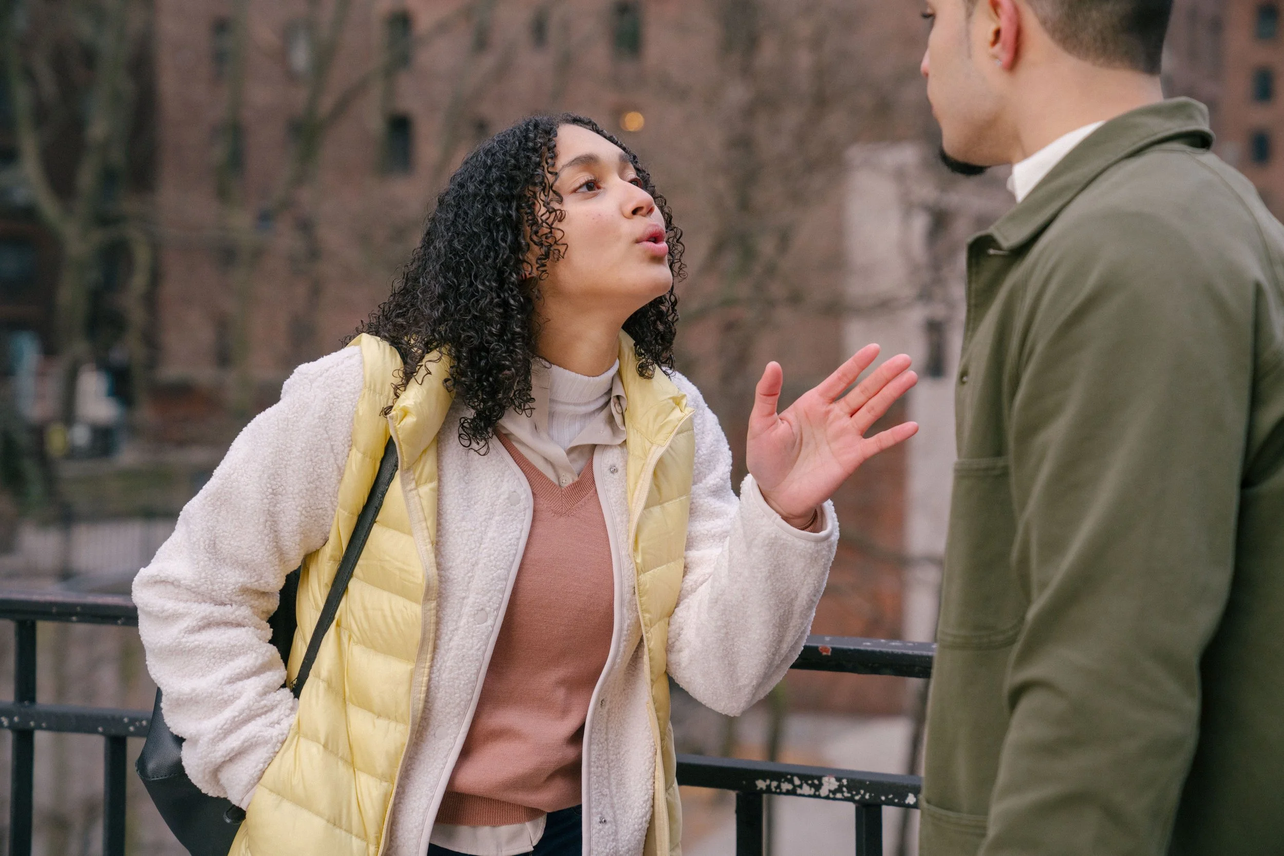 A woman with curly hair and a yellow jacket engaged in a heated conversation with a man outdoors on a city street, with trees and a brick building in the background.