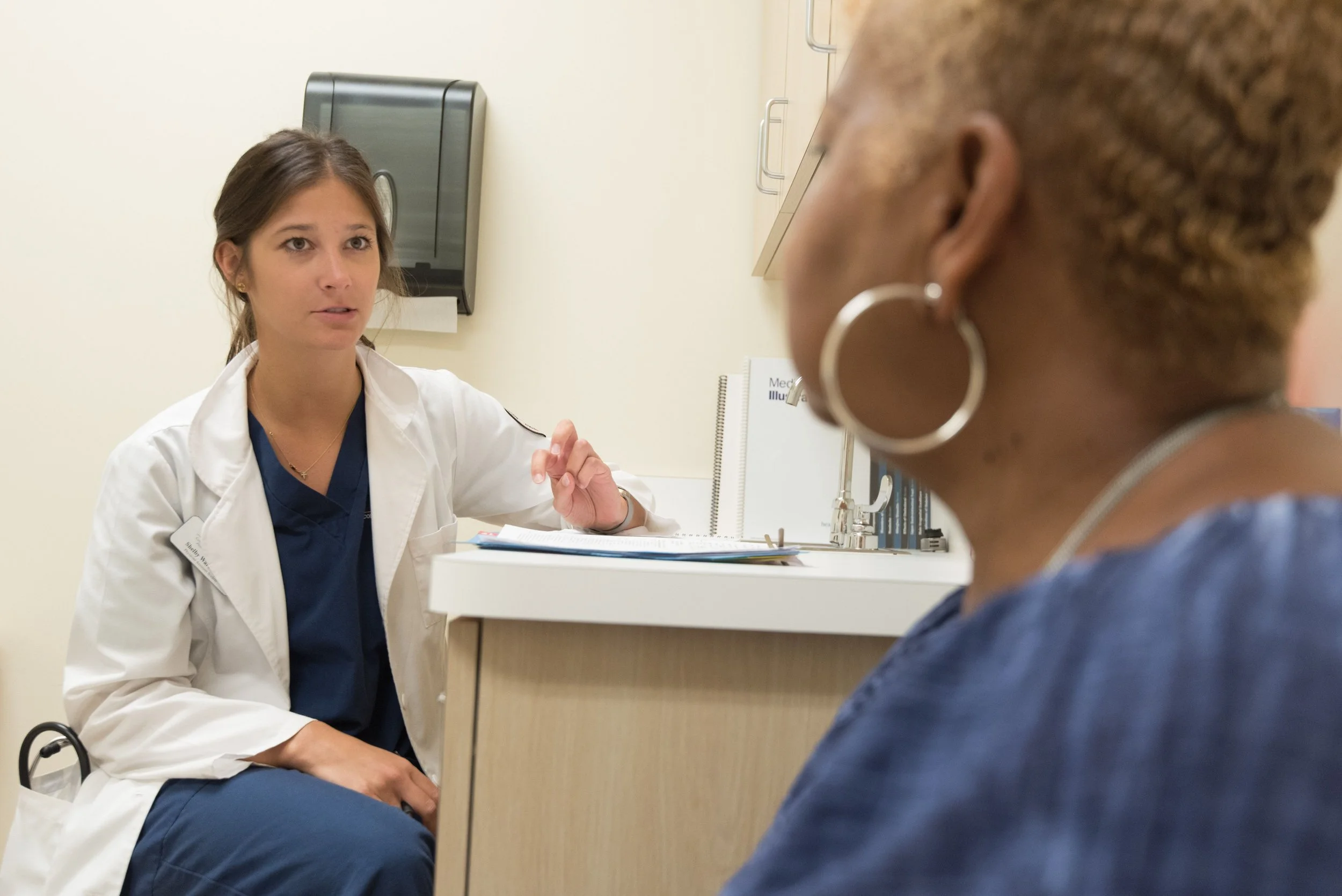 A female doctor in a white coat speaking with a patient in a medical office.