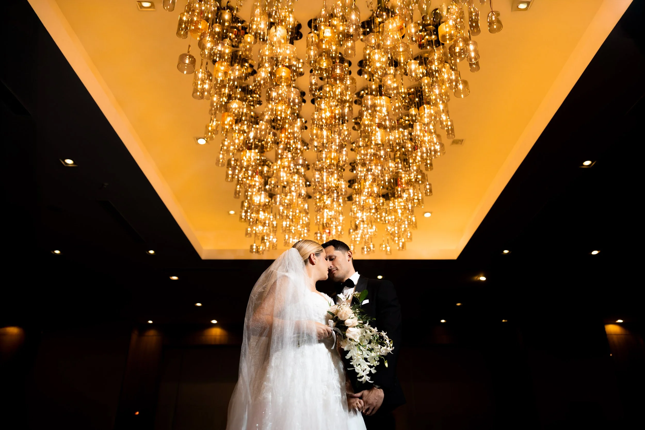Una pareja de recién casados en un acto en interiores, con iluminación cálida en el techo, la novia lleva un vestido de novia y velo, y el novio usa traje negro con pajarita, sosteniendo un ramo de flores blancas.