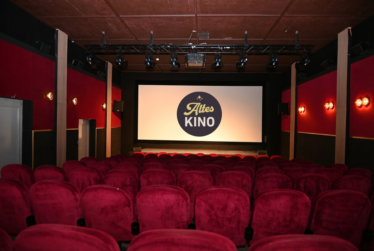 Ein leeres Kino mit roten, gepolsterten Sesseln und einer großen Leinwand, auf der das Logo "Altes Kino" zu sehen ist.